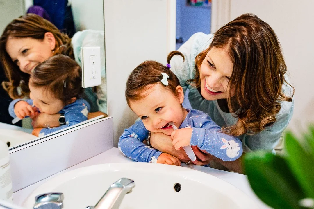 Mom helping her toddler brush her teeth in the bathroom mirror