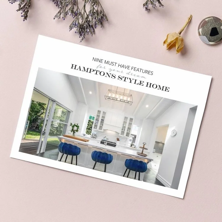 Cover page of a brochure or magazine featuring a modern white kitchen with blue bar stools and large windows, surrounded by flowers and decorative items on a pink surface.