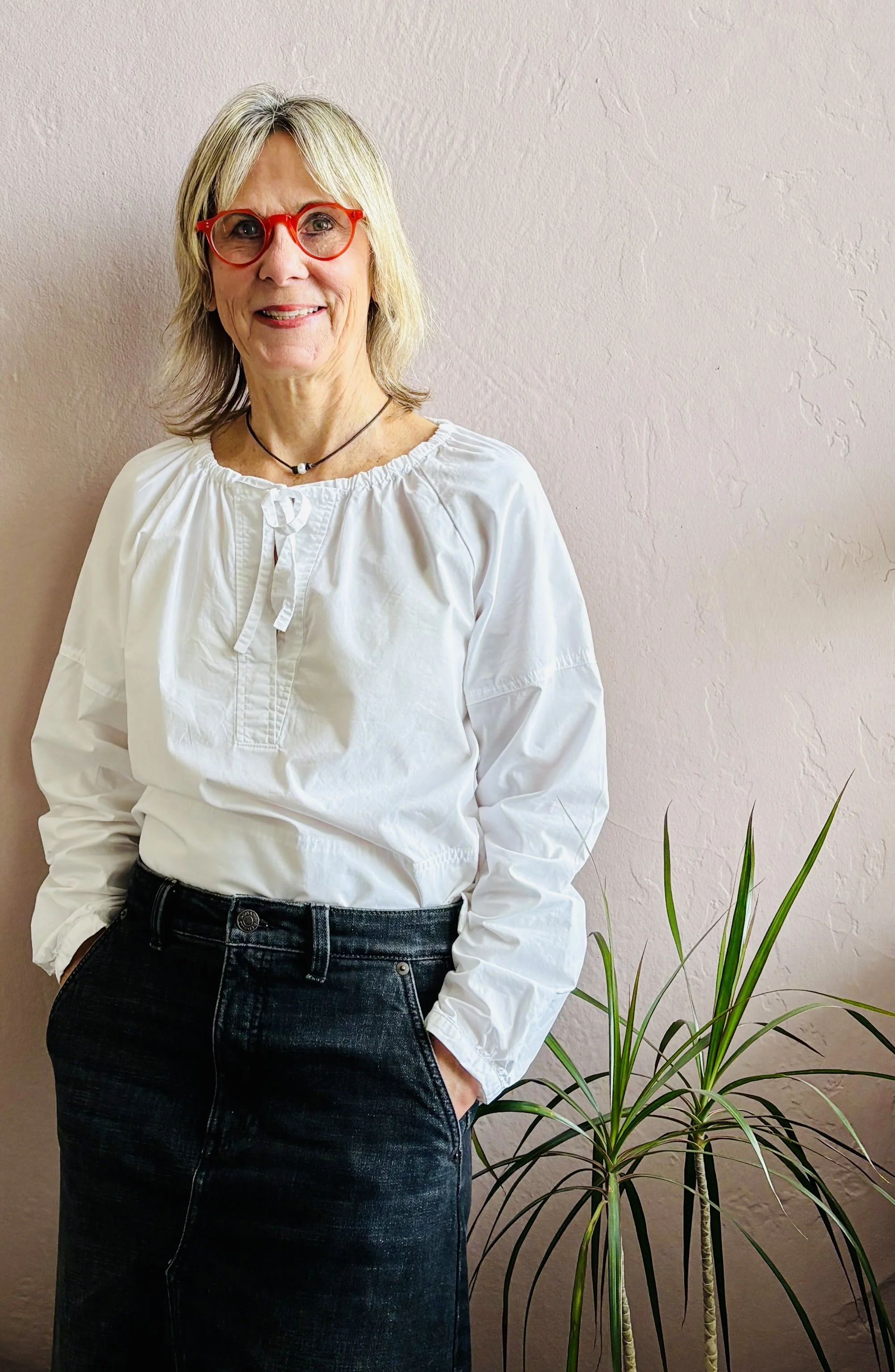 A woman with blonde hair, red glasses, and a white blouse standing next to a tall green plant against a light pink wall.