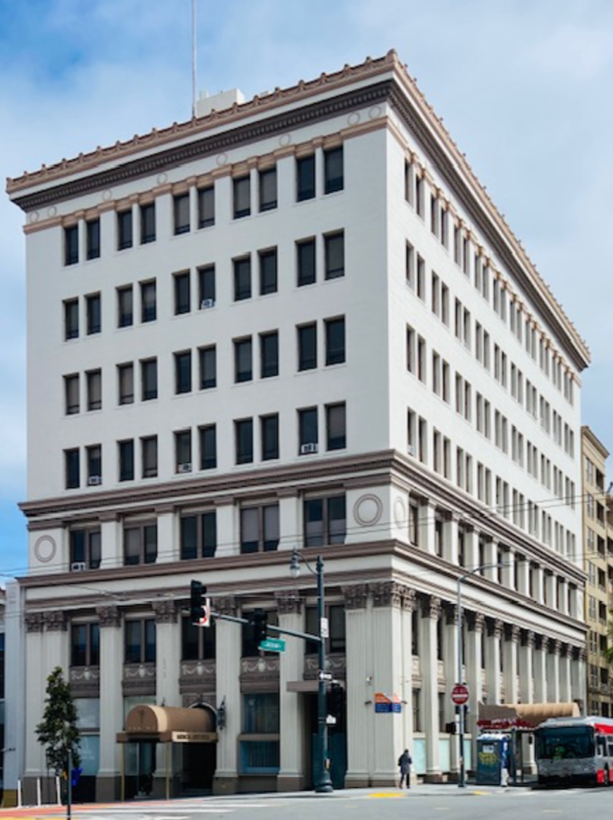 A multi-story white building with European architectural details, including columns and decorative trim, located on a city corner with sidewalk, traffic lights, and a bus nearby.