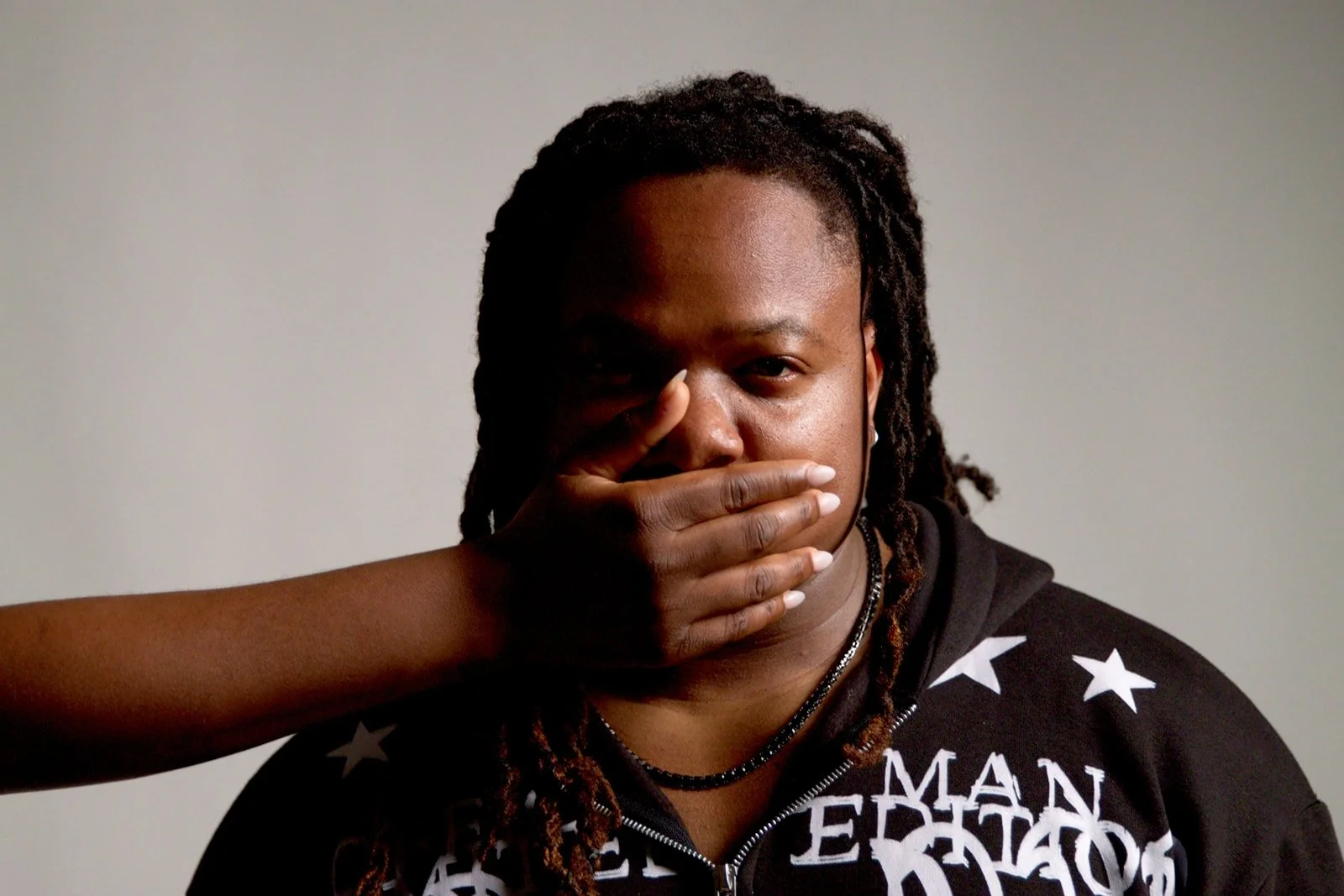 romenhermind with dreadlocks has their mouth covered by a hand, eyes looking directly at the camera, wearing a black shirt with white stars and text.