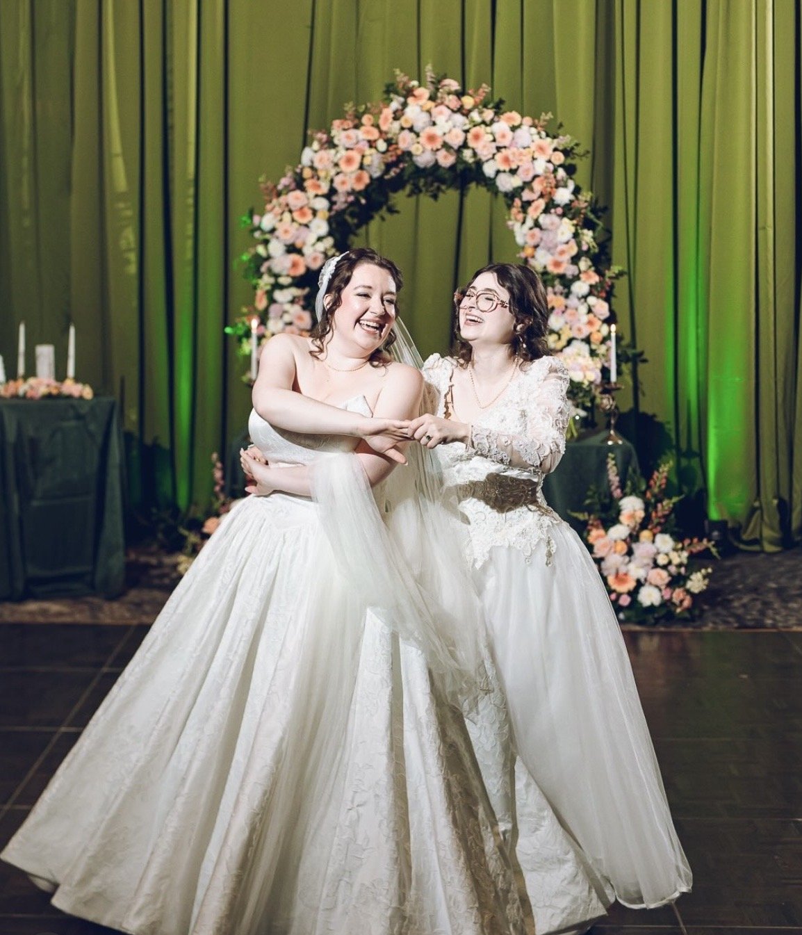 Two women in wedding dresses dancing and smiling at a wedding reception with a flower arch and green curtain backdrop.