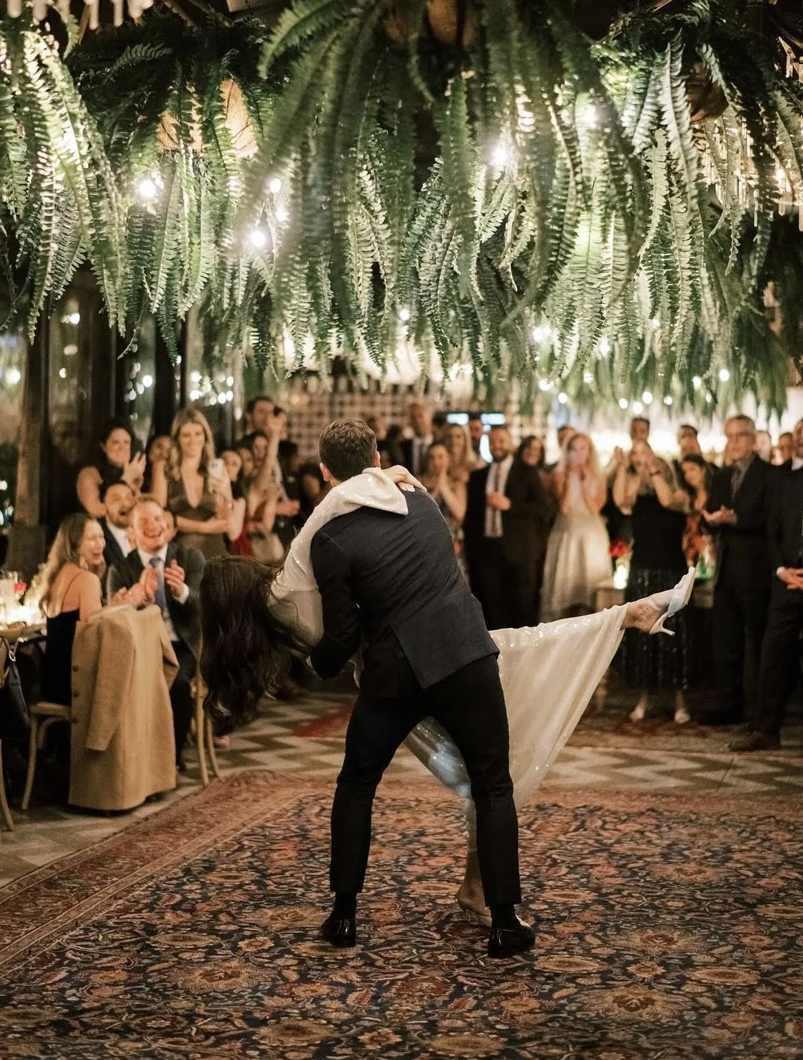 A couple dancing closely in the center of a lively celebration or wedding reception, with guests cheering and clapping around them under hanging greenery and string lights.