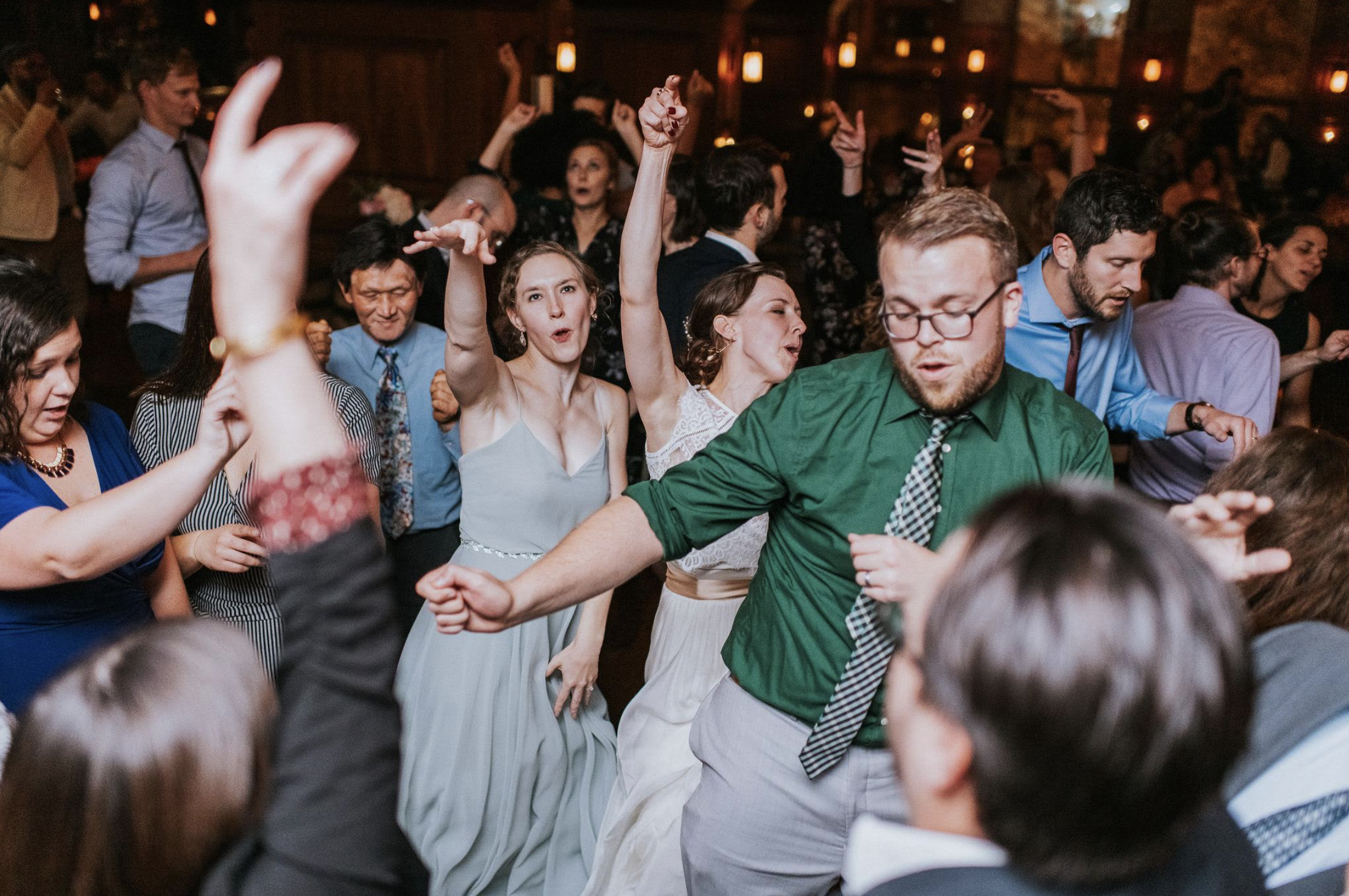 People dancing at a lively indoor party or celebration, with some raising arms in joy and movement, in a warmly lit venue.