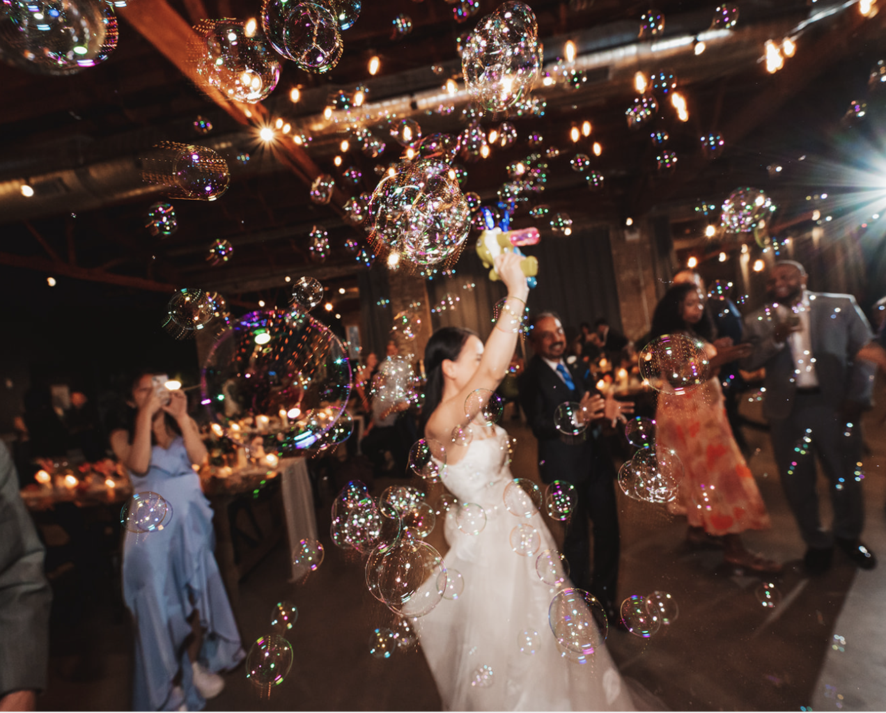 Wedding reception with bride in white dress holding bubble gun, surrounded by guests, bubbles in mid-air, warm lighting, wooden beams, and decorated venue.