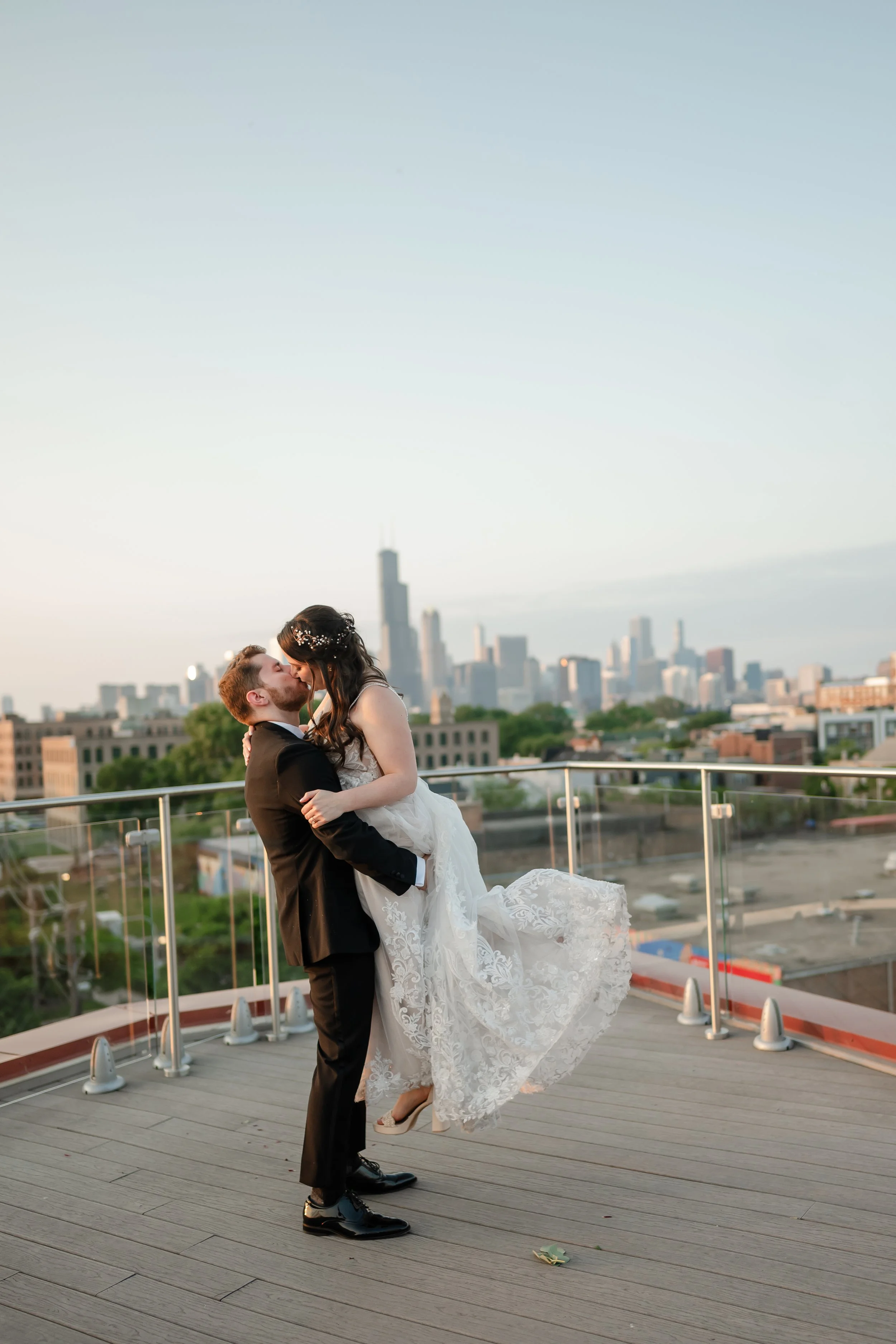 A bride and groom sharing a kiss on a rooftop with city skyline in the background during sunset. The groom is lifting the bride, who is wearing a white wedding dress, and they are embracing each other.