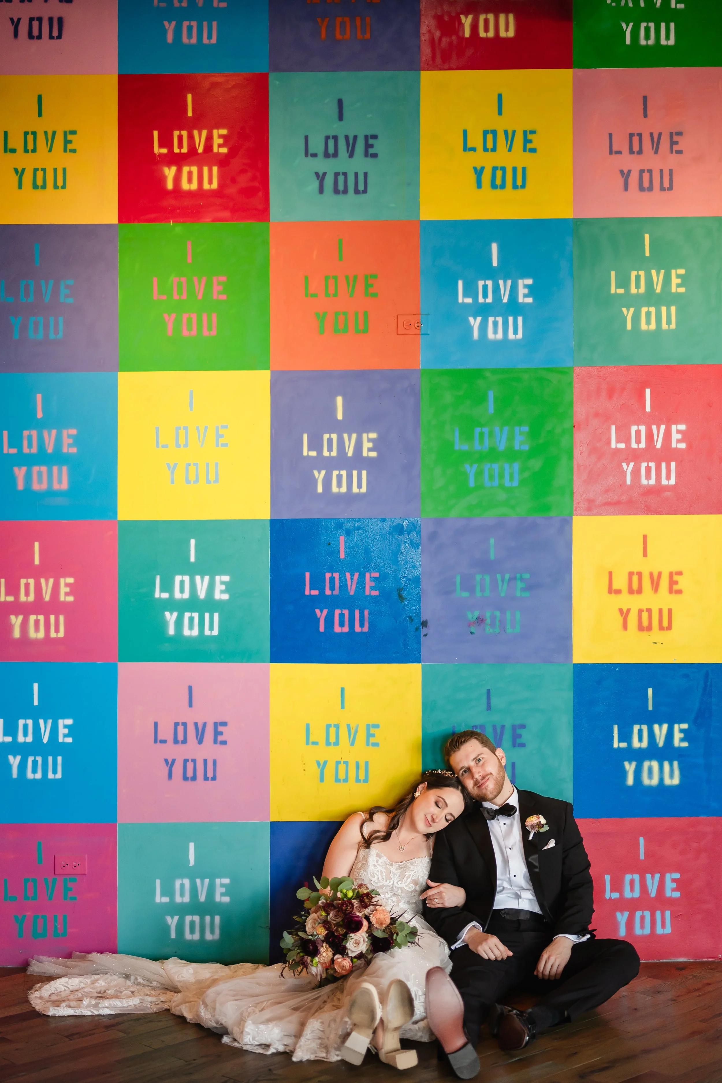 A bride and groom sitting on the floor in front of a colorful, patterned wall with repeated 'I love you' messages in bright colors. The bride has her head resting on the groom's shoulder, with her eyes closed, holding a bouquet. The groom is smiling 