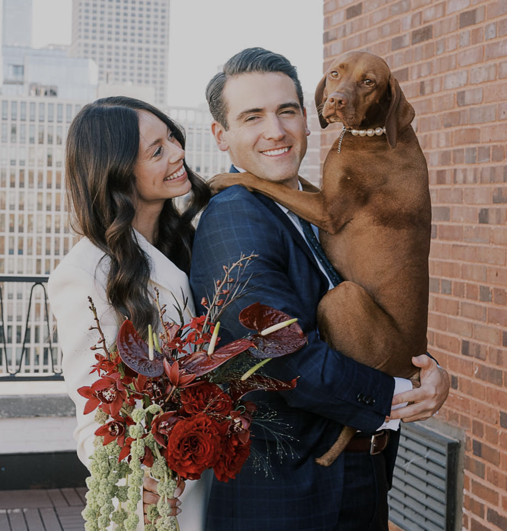 A happy couple with a brown dog, holding a bouquet of red flowers, standing on a rooftop with city buildings in the background.