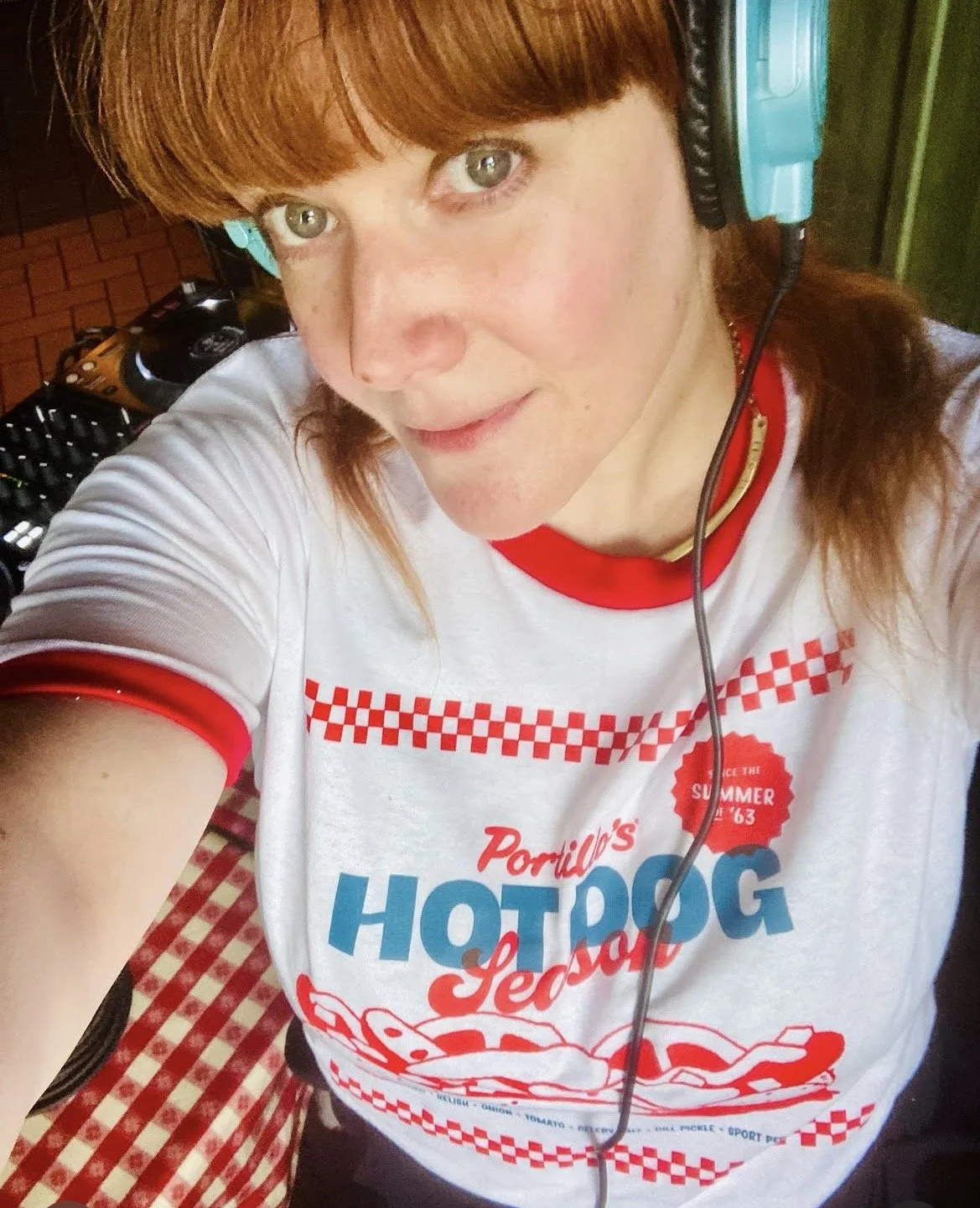 A woman with red hair wearing headphones and a white T-shirt with red and blue graphics, taking a selfie indoors.