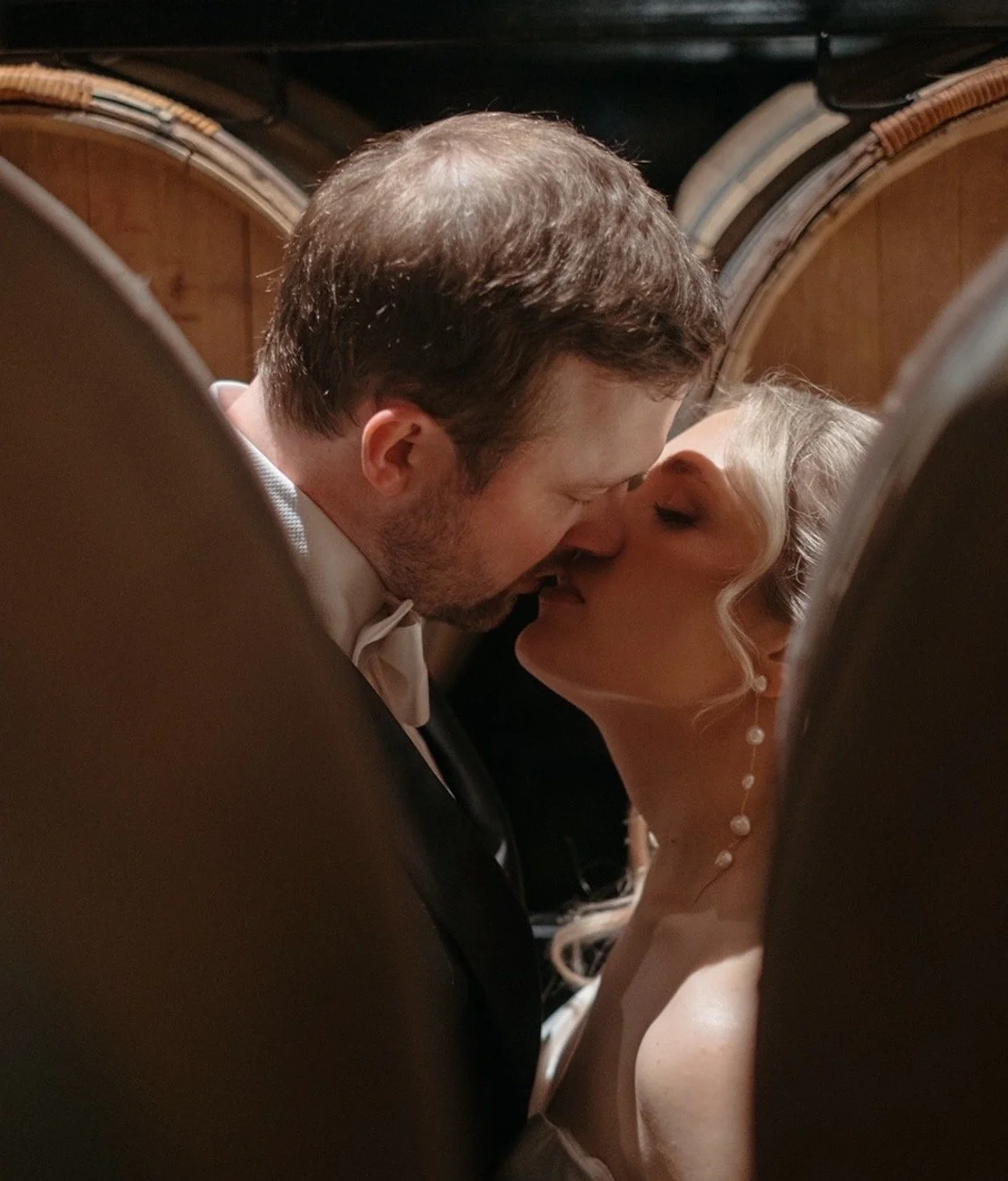 A bride and groom share a kiss in a close-up shot framed by pews, with warm lighting and wooden background.