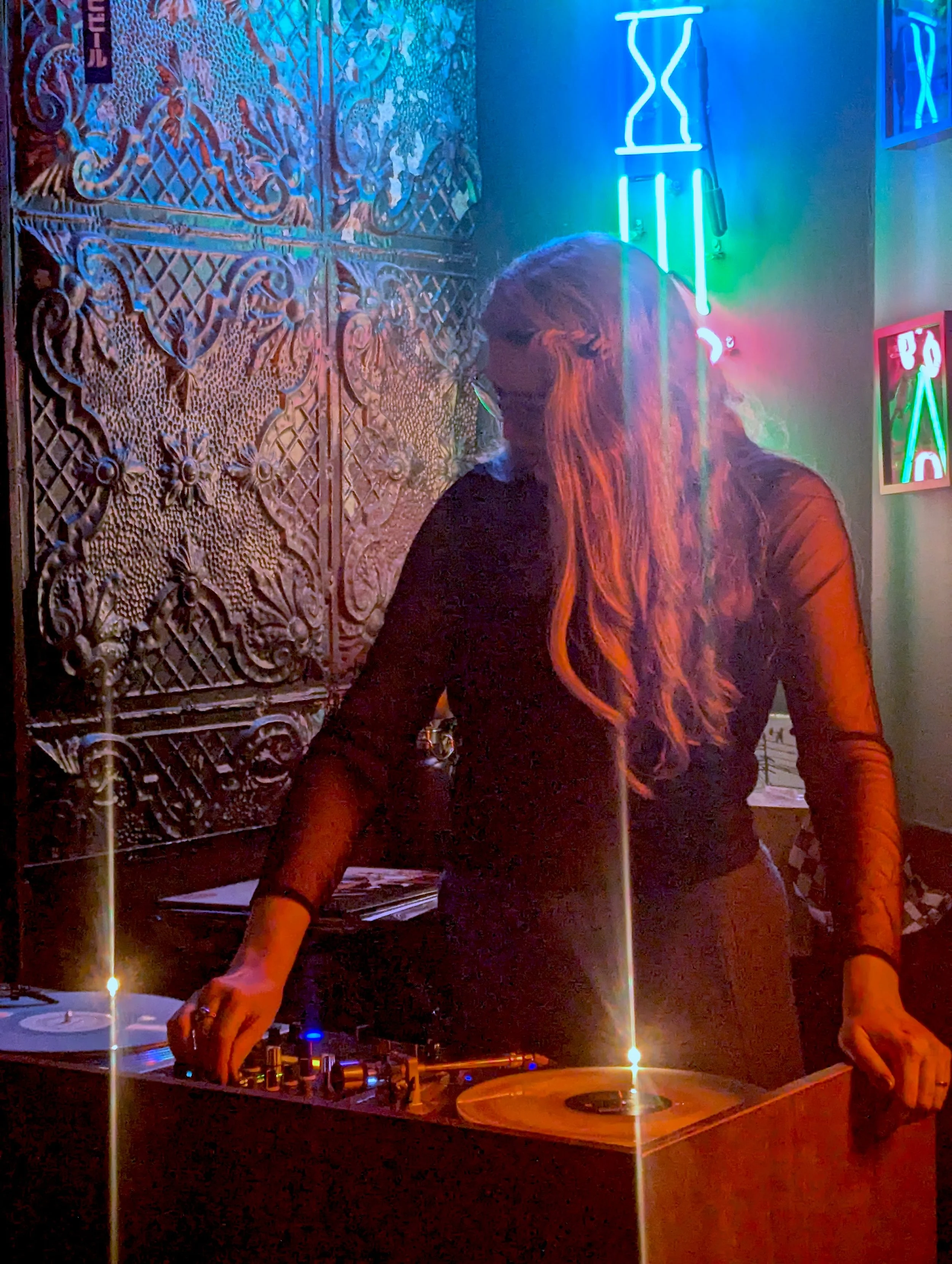 A DJ with long hair operating turntables and a mixer in a dimly lit room with decorative metal wall art and neon blue, green, and pink lighting.