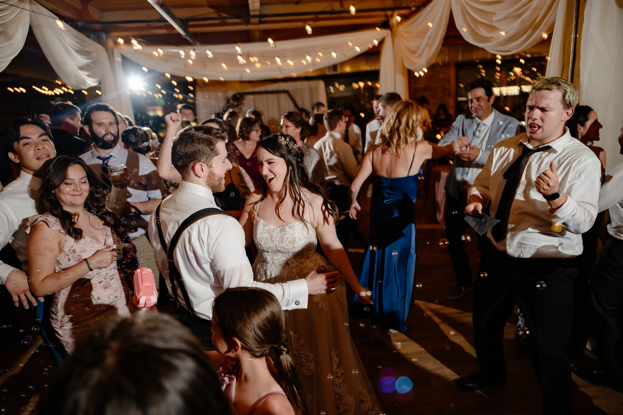 People dancing and celebrating at a wedding reception with string lights and draped fabric decorations.