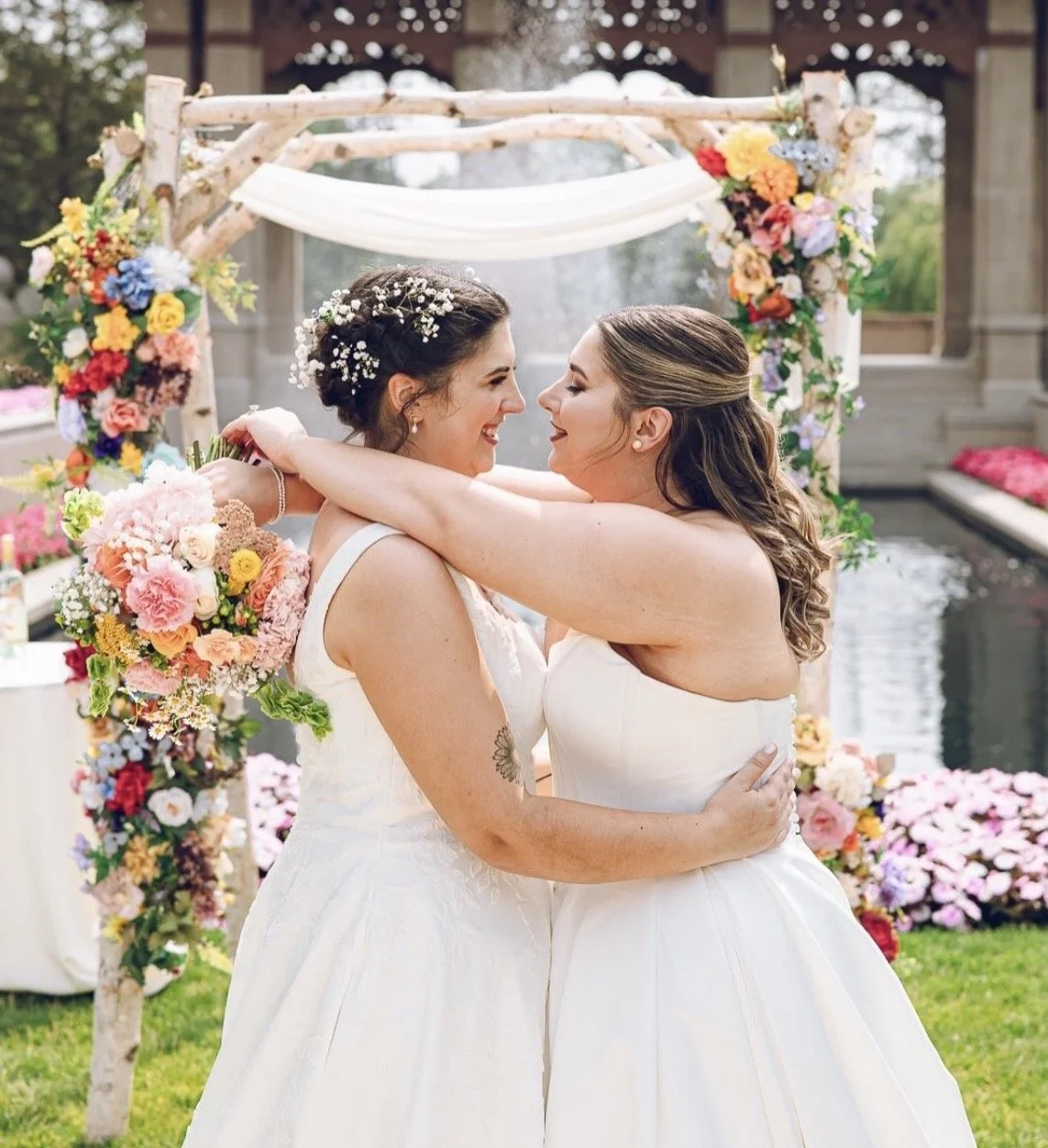Two women in wedding dresses embrace smiling at each other during a wedding ceremony outdoors with a floral arch in the background.