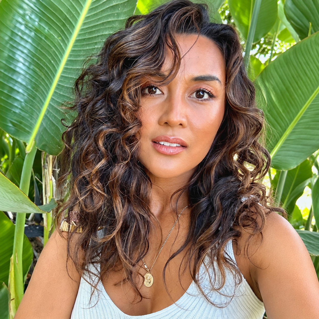A woman with curly brown hair takes a selfie outdoors amid large green tropical leaves, wearing a white sleeveless top and a gold necklace.