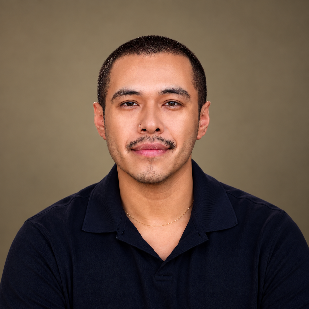 Portrait of a young man with short dark hair, a slight mustache, and light brown skin, wearing a black polo shirt and a delicate gold chain, smiling softly against a neutral background.