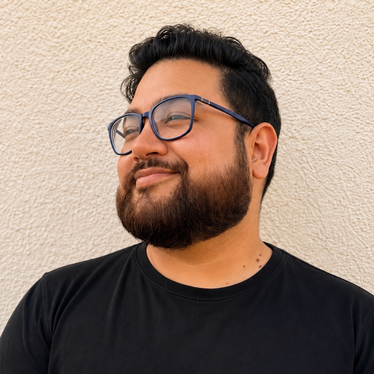 A man with glasses and a beard smiling and looking to his left against a beige textured wall.