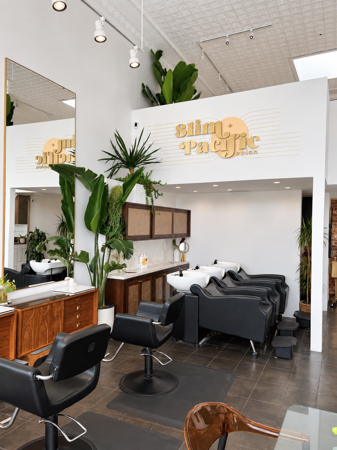 Interior of a modern hair salon with black styling chairs, shampoo stations, large mirror, plants, and a sign reading 'slim + pacific salon' on a white wall.