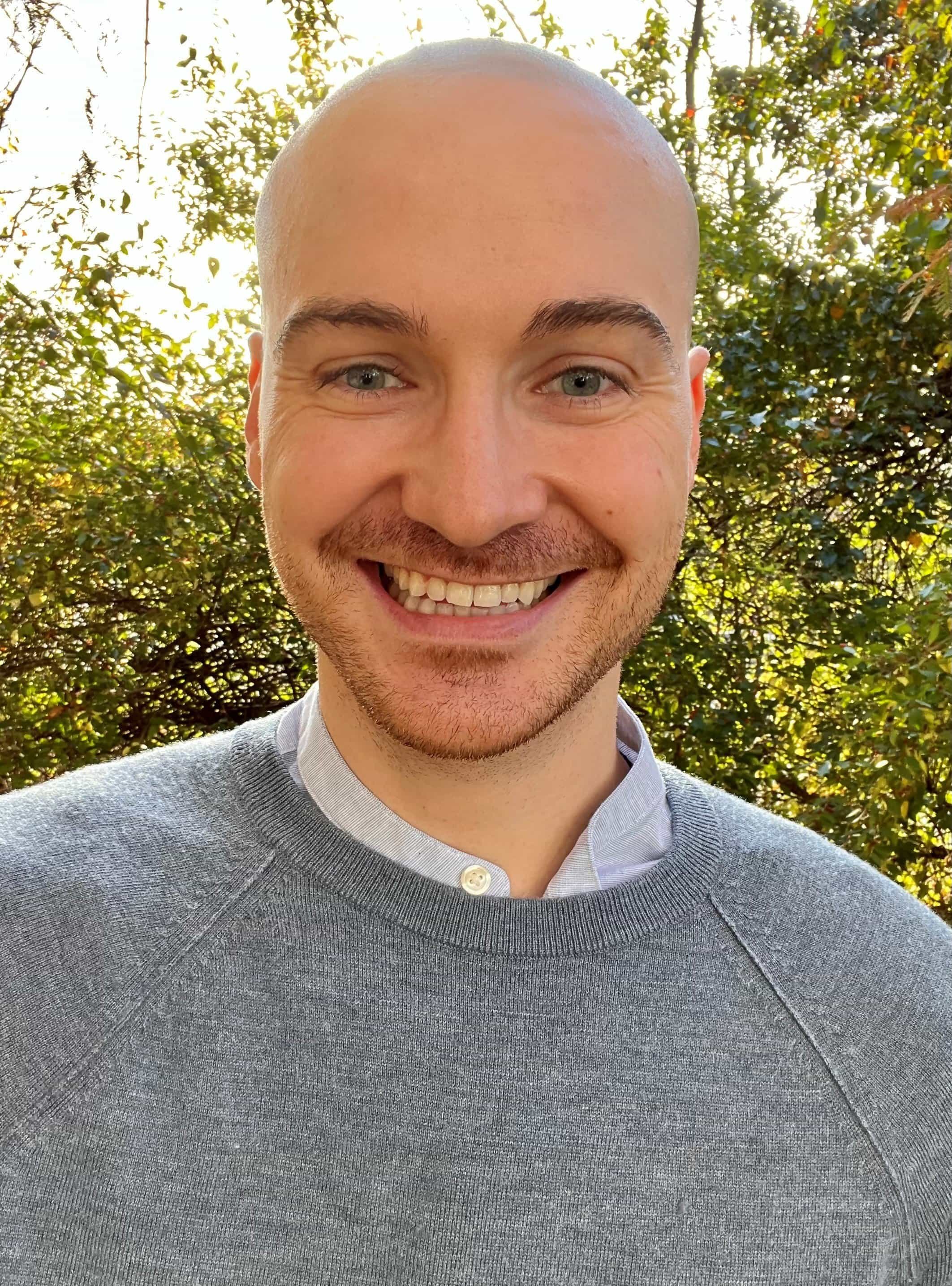 A smiling man with a shaved head and light beard outdoors, with green foliage and sunlight in the background.