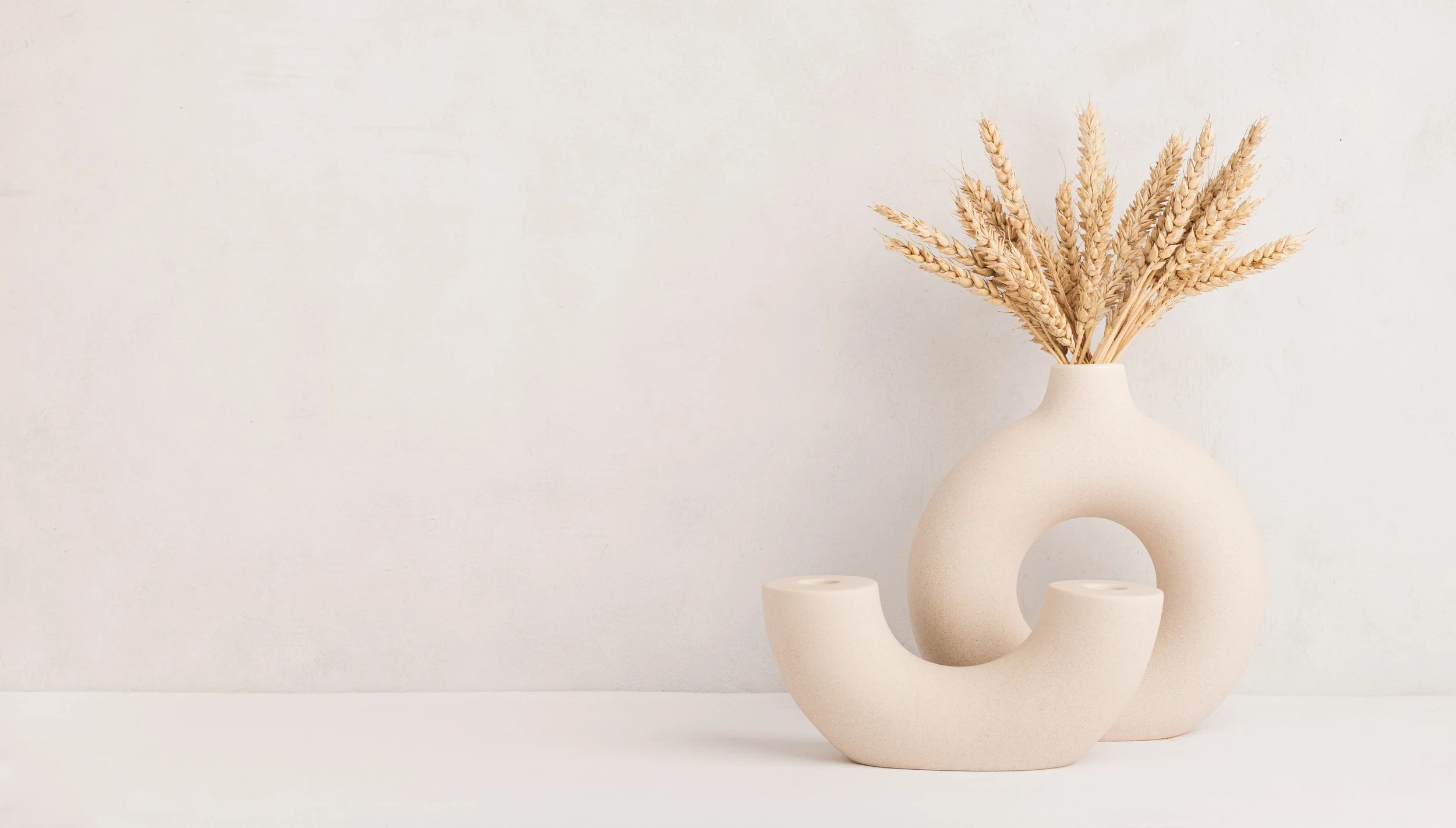 Minimalist beige ceramic vases with dried wheat stalks on a white surface against a white wall