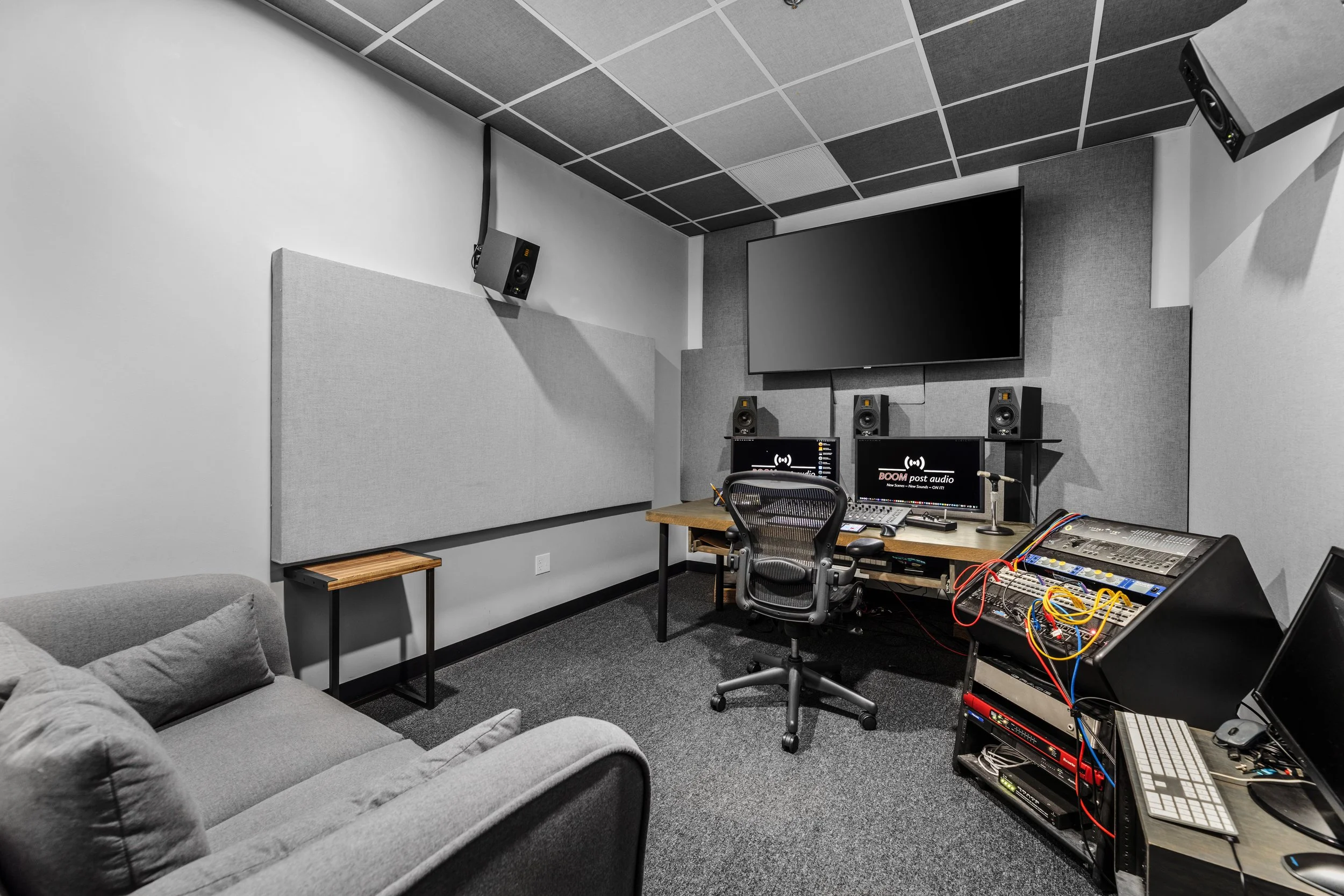 A professional recording or podcast studio featuring a large wall-mounted monitor, multiple studio speakers, a computer with audio editing software, a mixing console, a microphone, and various audio equipment in a gray and black color scheme.