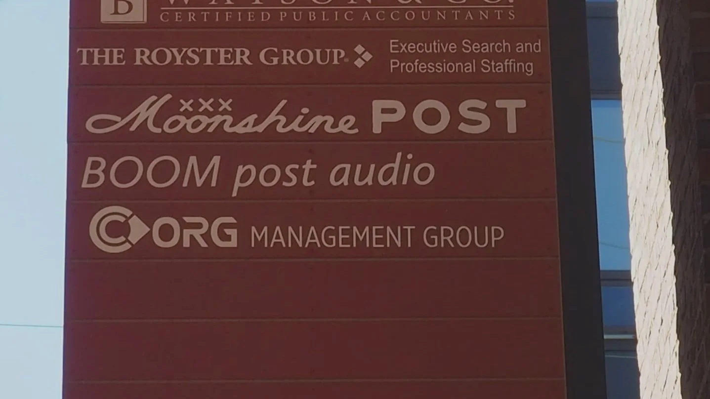A signboard displaying multiple business listings, including The Royster Group, Moonshine POST, BOOM post audio, and ORG Management Group.