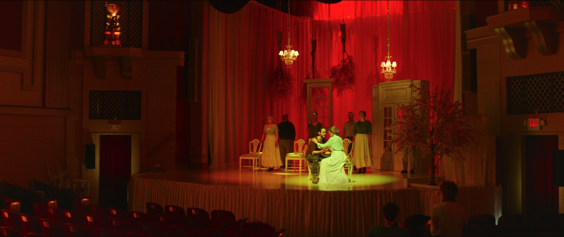 Theatrical performance on stage with actors, set decorated with red curtains, chandeliers, and furniture, with audience seating in the foreground.