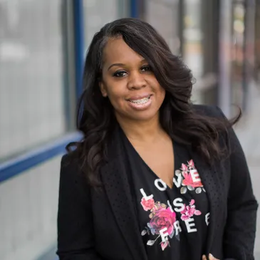 Circle image of a Black woman wearing a black tshirt with roses that says "Love is Free" and a black blazer. She has long black hair and is looking at the camera.