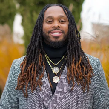 Circle image of a Black man with a beard smiling at the camera. He has long black and highlighted dreadlocks and is wearing two pendant necklaces and a gray tweed blazer