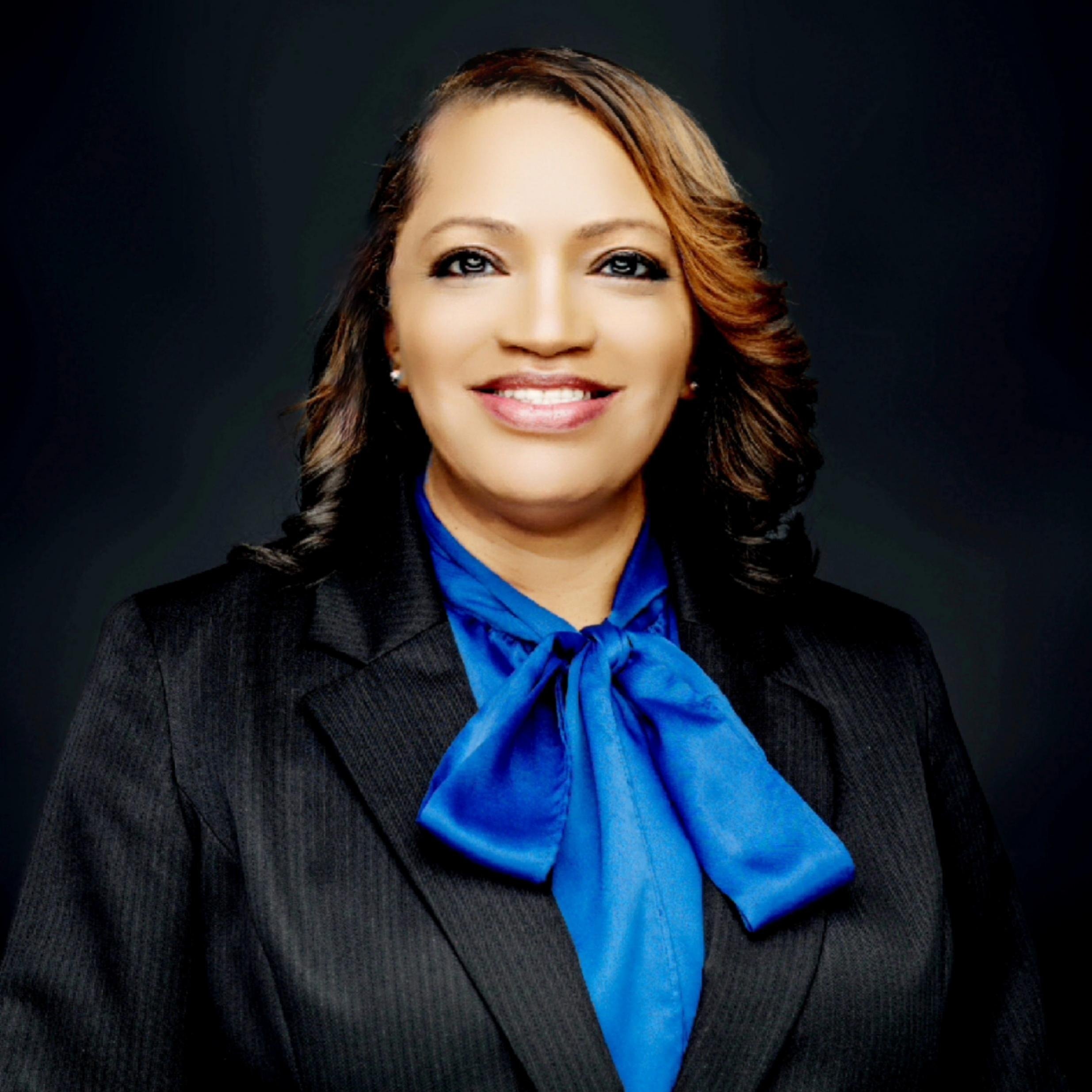 Black woman with brown hair looking at the camera and smiling, wearing a black jacket, white tshirt and turquoise necklace. Smiling and wearing a blue blouse and black blazer.