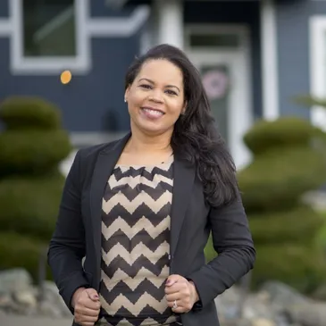 Circle image of Black woman smiling at the camera with long black hair and wearing a black blazer and black and white striped blouse. She is wearing earrings and a ring.