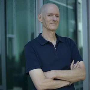 A man with a shaved head, wearing a navy blue polo shirt, standing outdoors in front of a modern glass building, with his arms crossed and a slight smile.