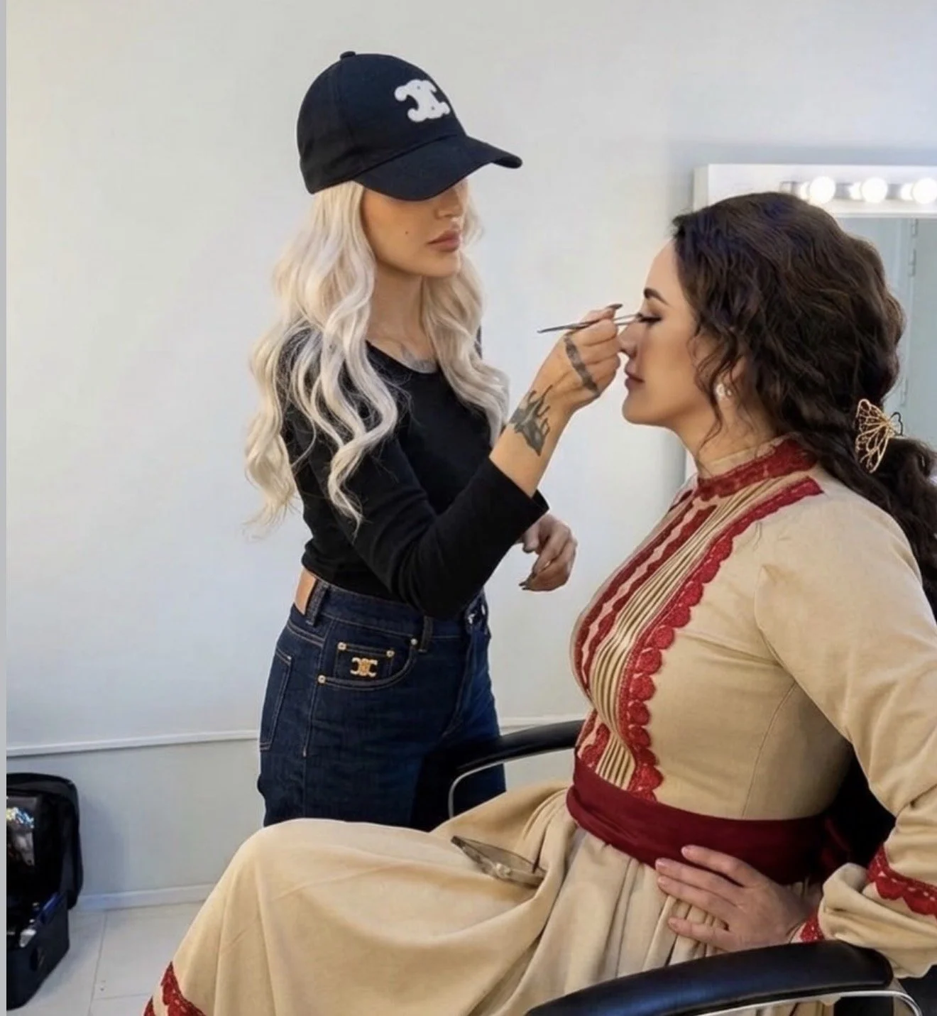 A makeup artist applies eye makeup to a woman sitting in a chair. The makeup artist has long, wavy blonde hair, is wearing a black cap, and a black long-sleeve shirt. The seated woman has dark, curly hair styled with a butterfly hair clip, and is wea