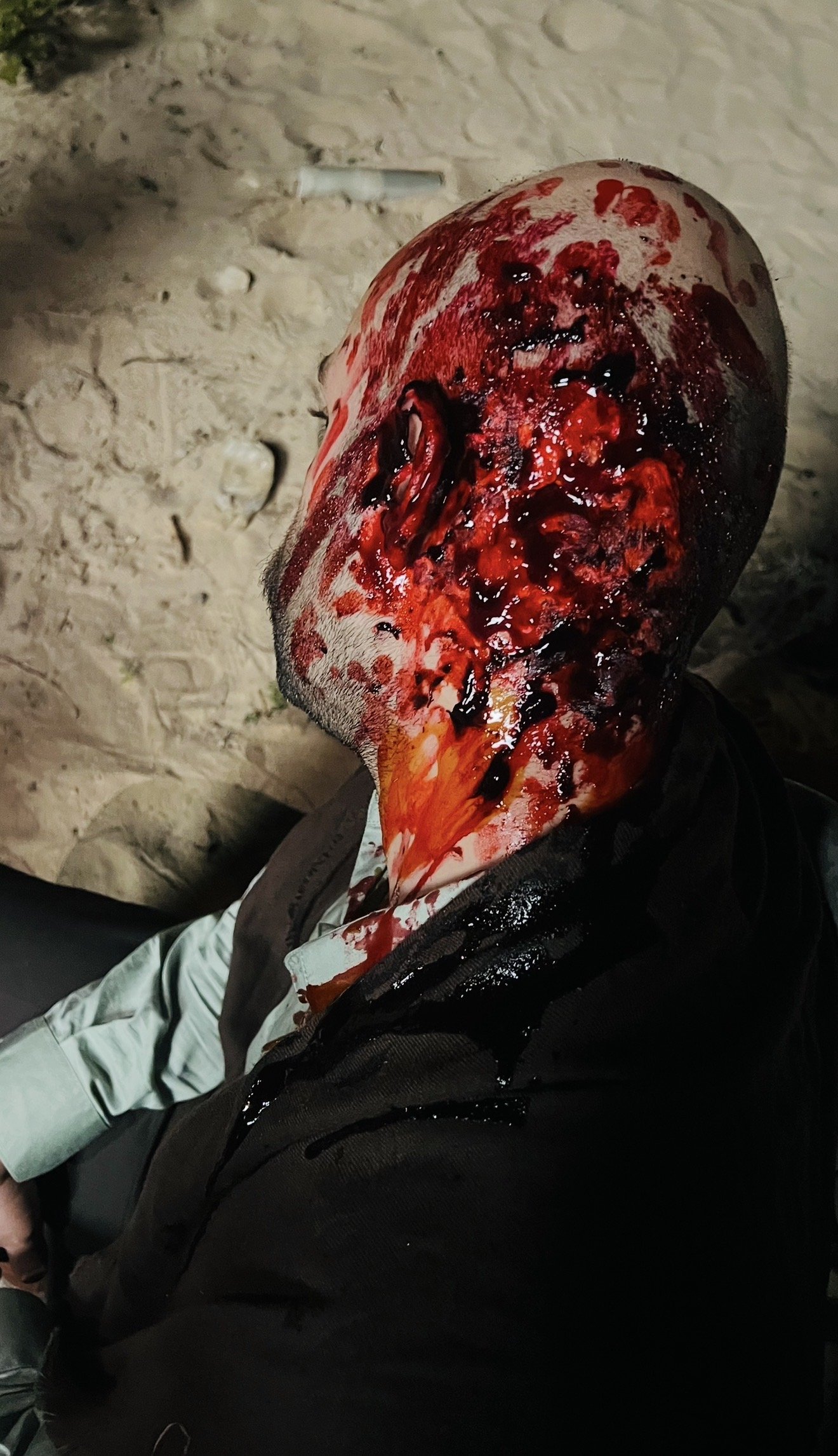 A person's head with severe injuries, blood, and open wounds, lying on the ground with sand or dirt around.