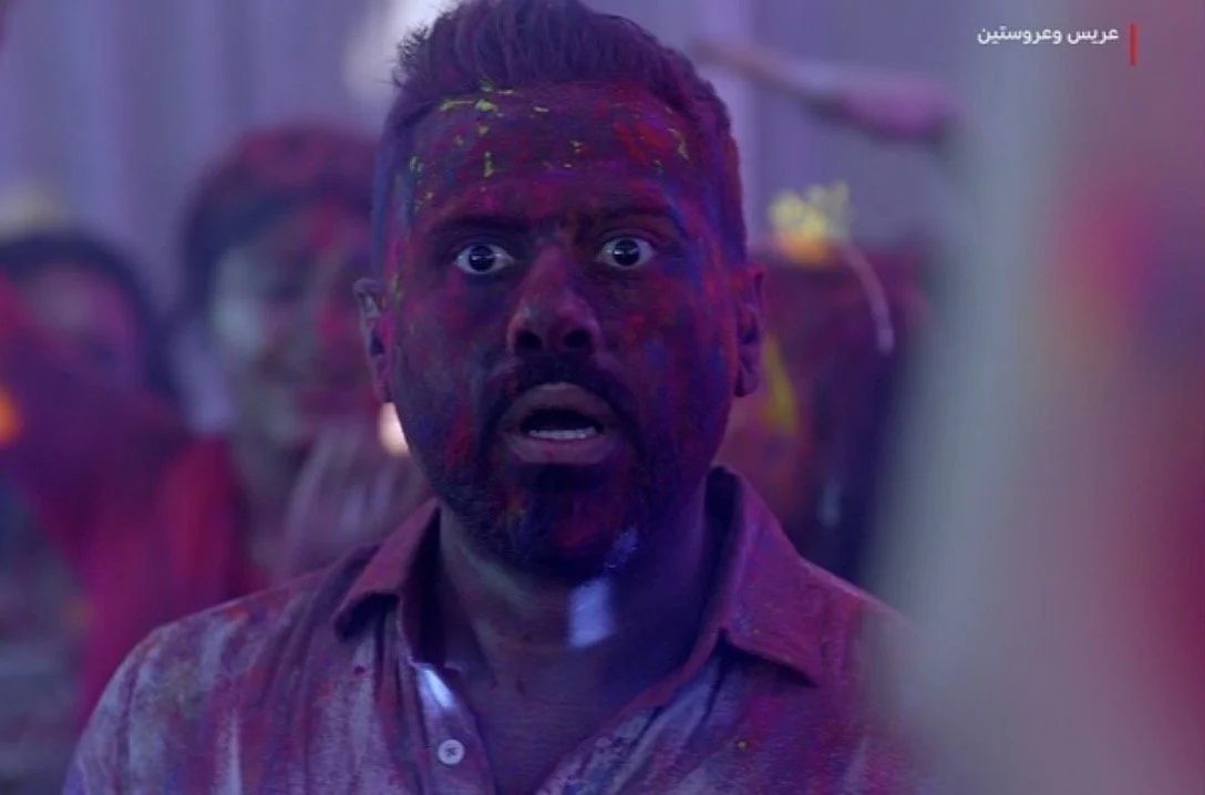 Man with face and clothing covered in colorful powder, looking surprised or shocked, at a celebration or festival during the festival of Holi.