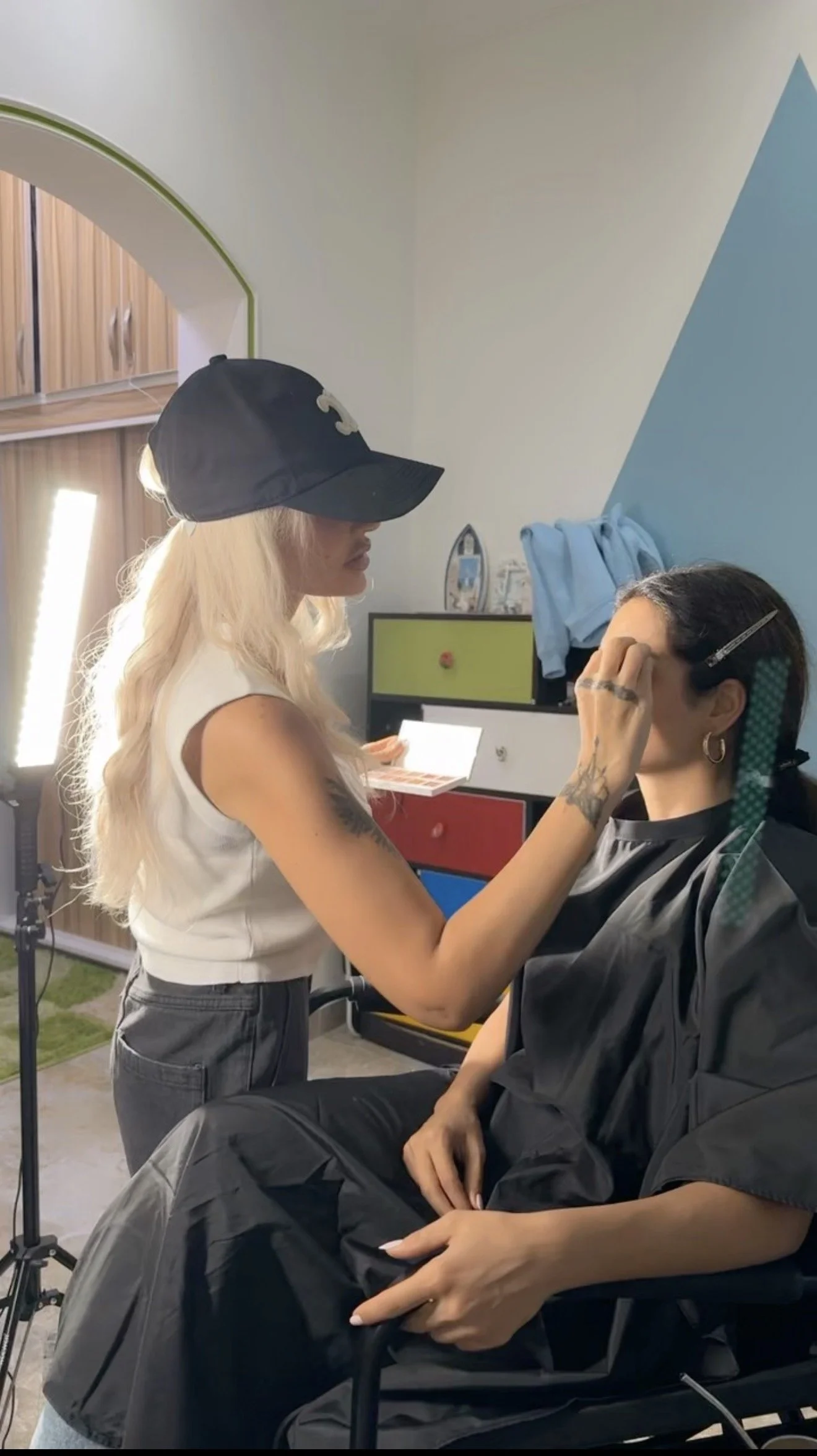 A woman with long blonde hair, wearing a black baseball cap and white sleeveless top, is applying makeup or face paint to a seated woman with dark hair, who is wearing a black cape and seated in a salon or studio setting.