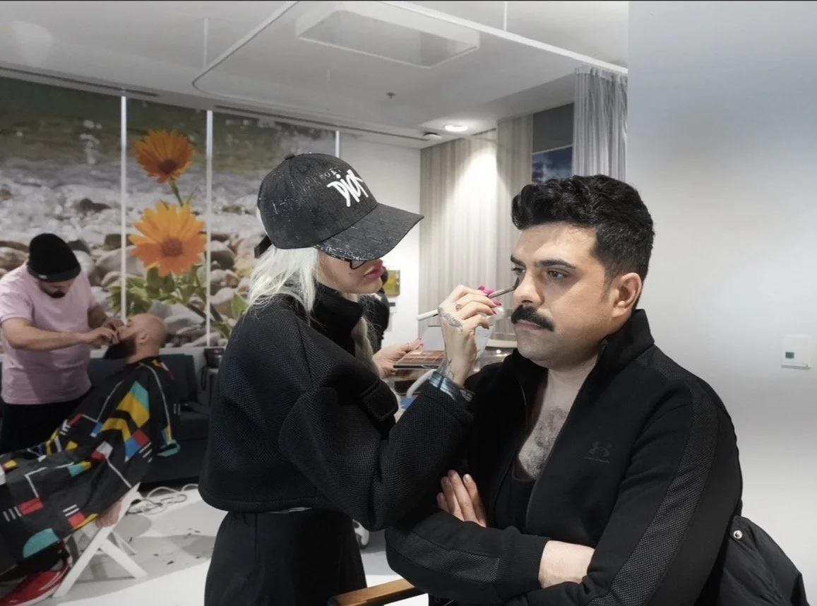 A woman applying makeup on a man with a mustache while another person is in the background doing hair.