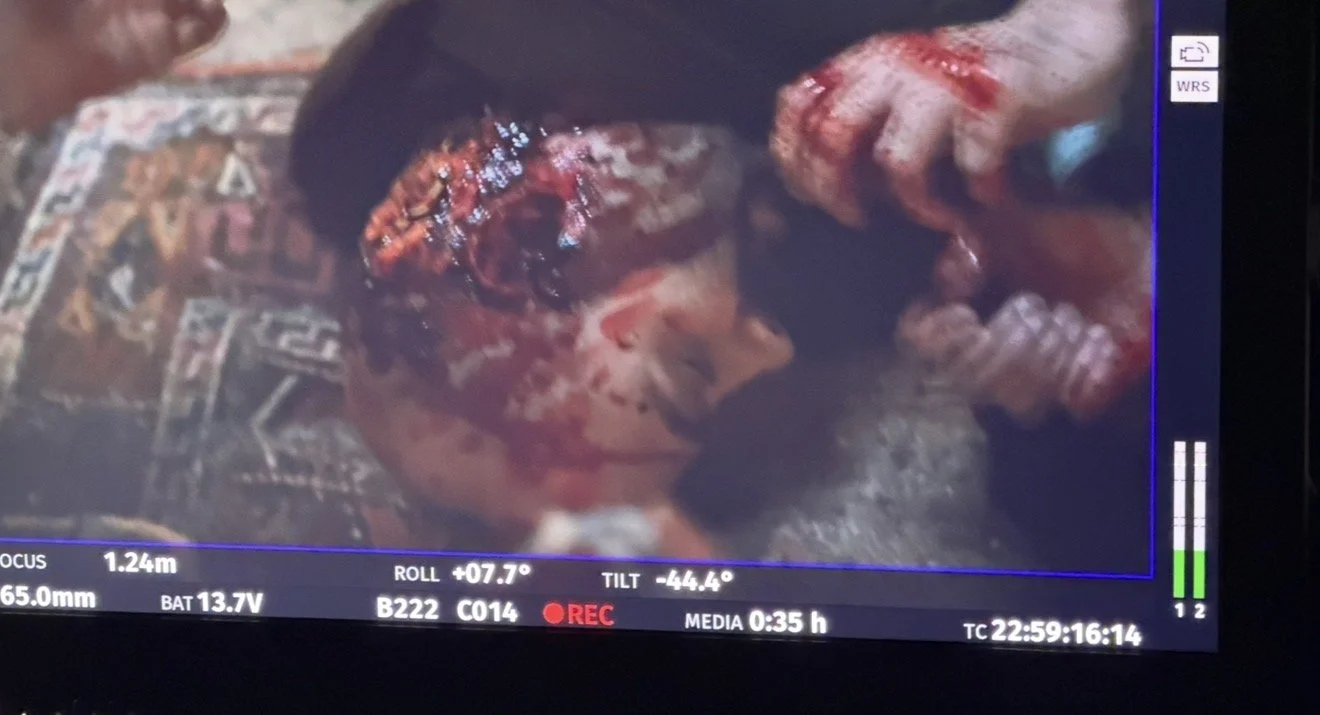 A person with short dark hair lies unconscious with blood on their face, while a hand appears to be holding their head. The scene is bloodstained and appears to be from a film or video recording, with camera details and recording time displayed on th