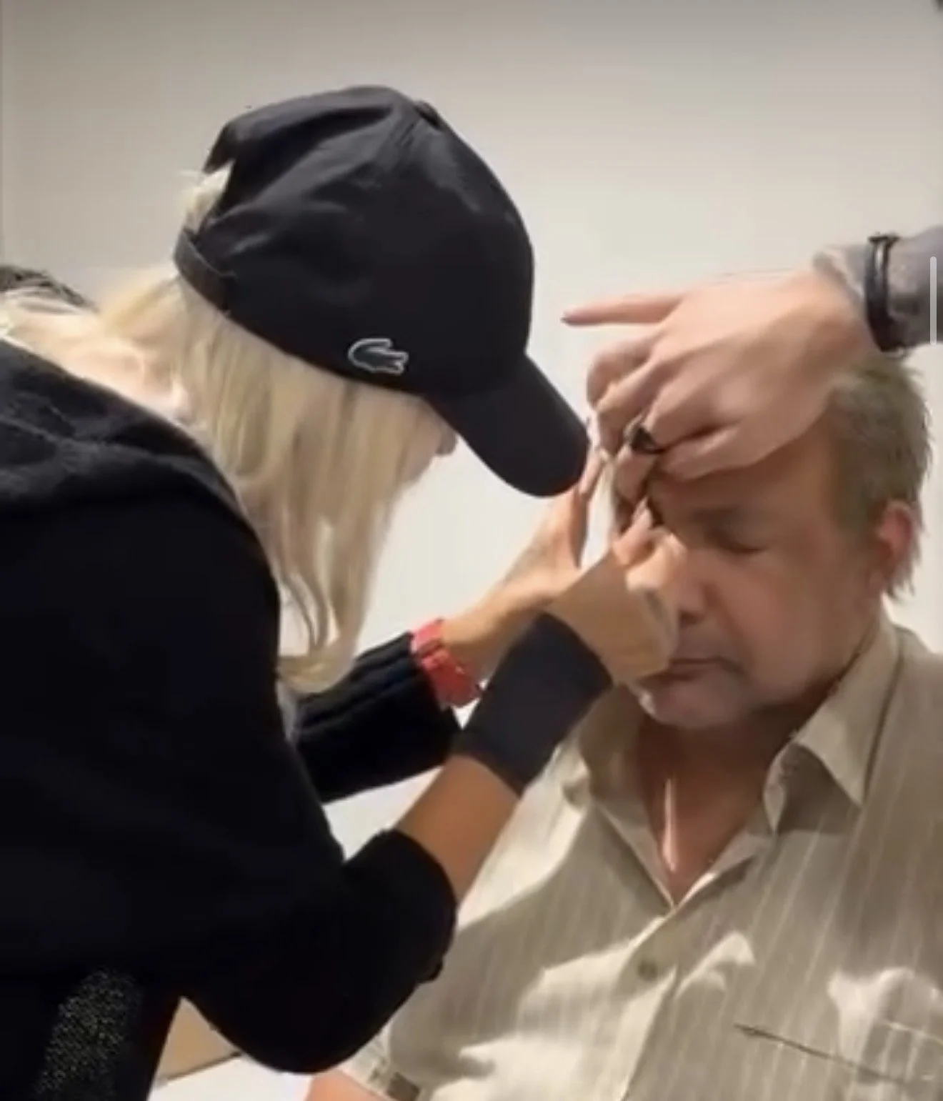 A woman with blonde hair wearing a black cap and black long-sleeve shirt is applying makeup or face paint to a man's forehead. The man has blonde hair, a beige shirt with vertical stripes, and is sitting with his eyes closed.