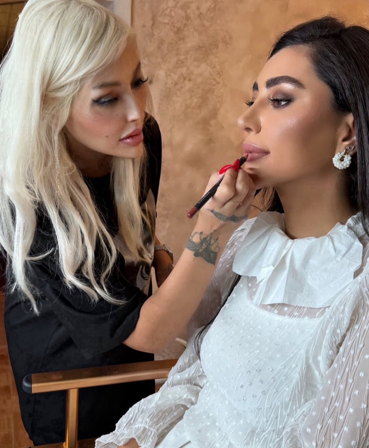 A makeup artist applying lipstick to a woman with dark hair and earrings, sitting in a chair with a white textured dress.