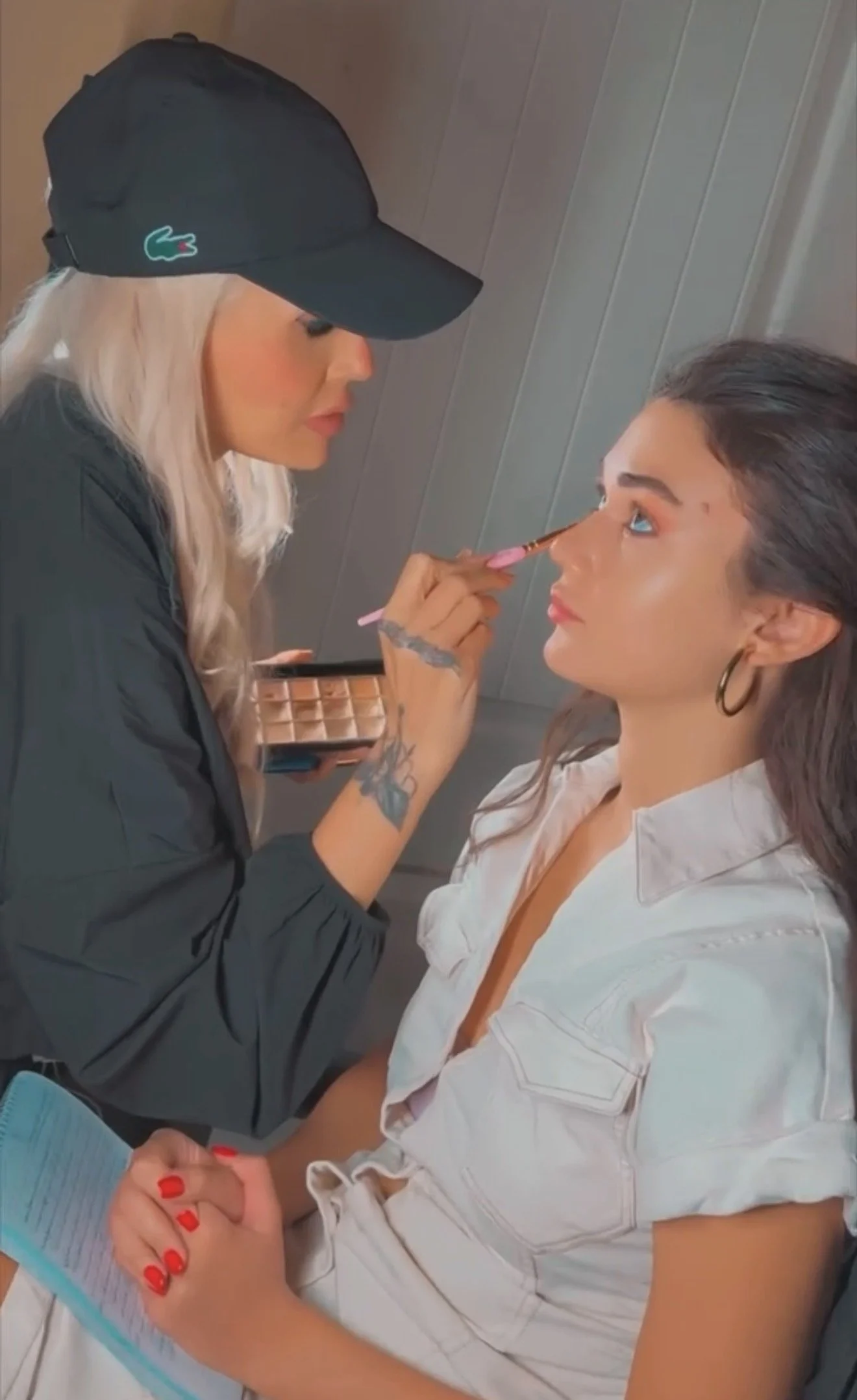 A woman with dark hair and hoop earrings sits while another woman with long blonde hair, wearing a black cap and jacket, applies makeup to her nose with a brush. The woman receiving makeup has a light-colored shirt and is holding a notebook.