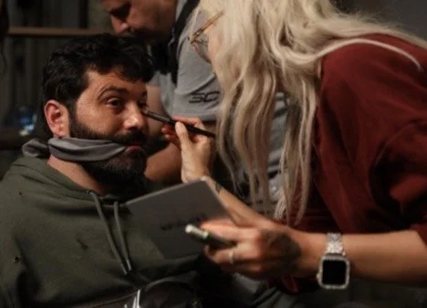 A woman with blonde hair applying makeup or face paint to a man's face, who is seated and holding a tablet, with a man in the background observing.