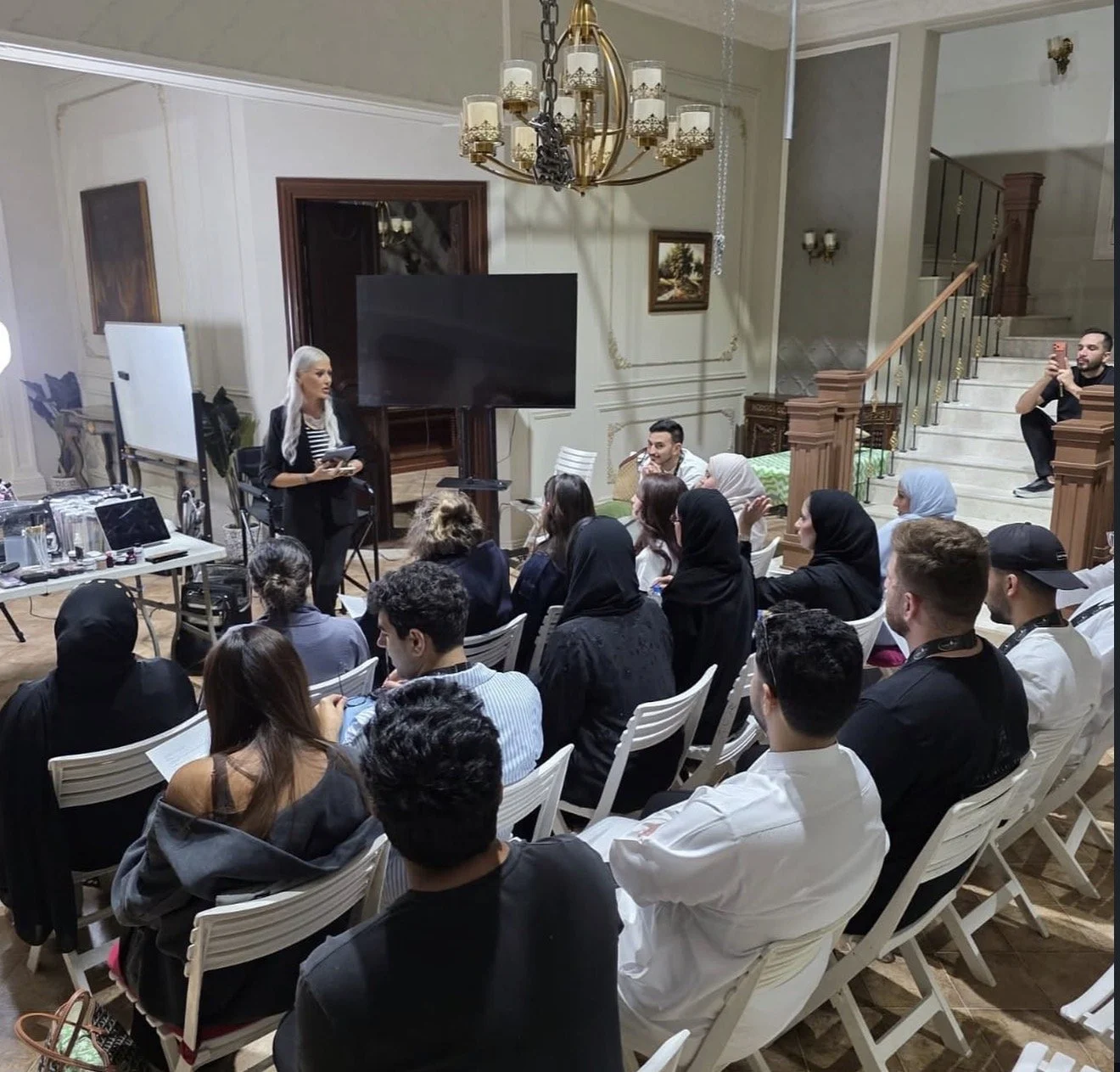 A woman standing and speaking to a seated audience in a spacious, elegantly decorated room with a chandelier and staircase.