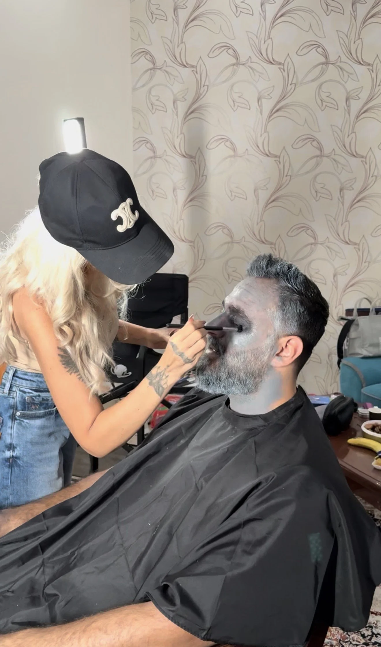 A man with gray hair and beard getting makeup applied to his face by a woman with long blonde hair and a black cap. The woman is wearing a black cap and jeans, and the man is draped in a black salon cape. They are in a room with patterned wallpaper.