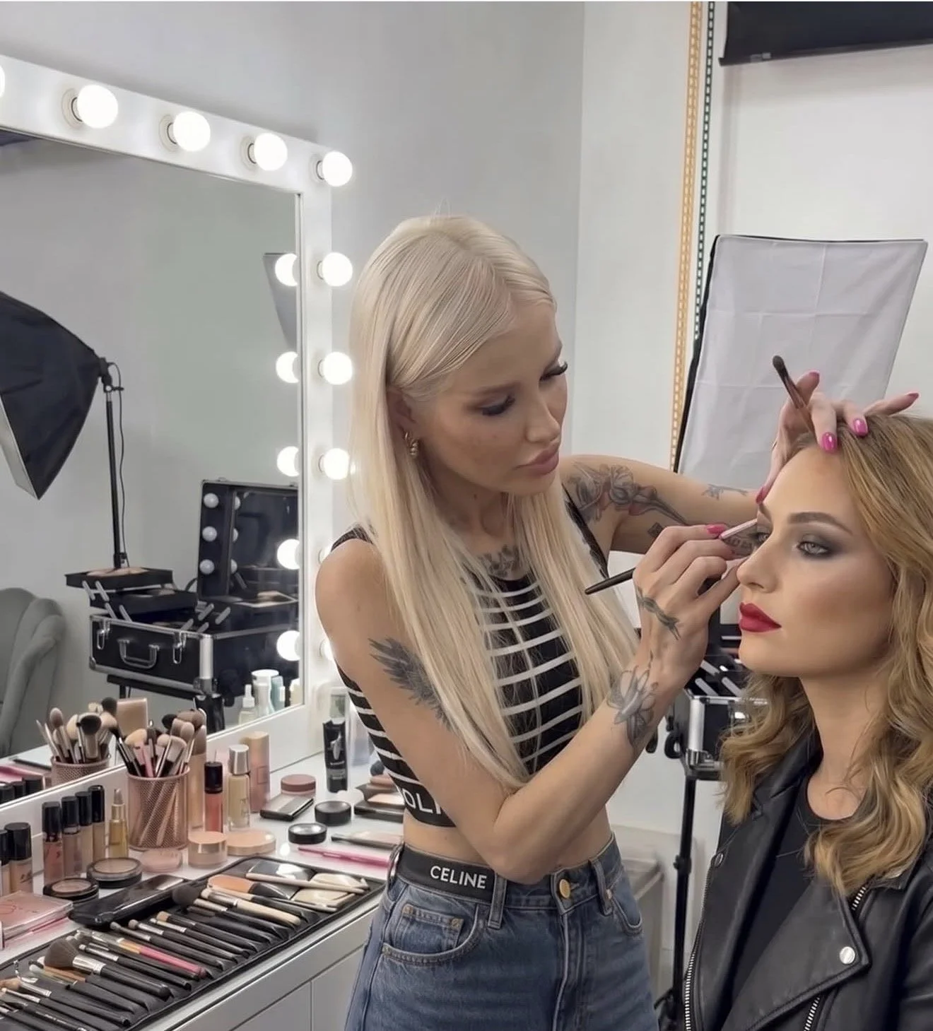 A makeup artist applying makeup to a woman in a professional makeup studio. The studio has a large mirror surrounded by bright lights and a table filled with various makeup tools and products.
