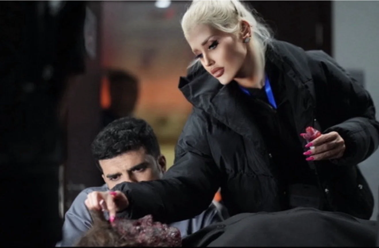 A woman in a black jacket leaning over a man with a gunshot wound, examining his injury, while a person appears to be lying down on a table or surface, in an indoor setting.