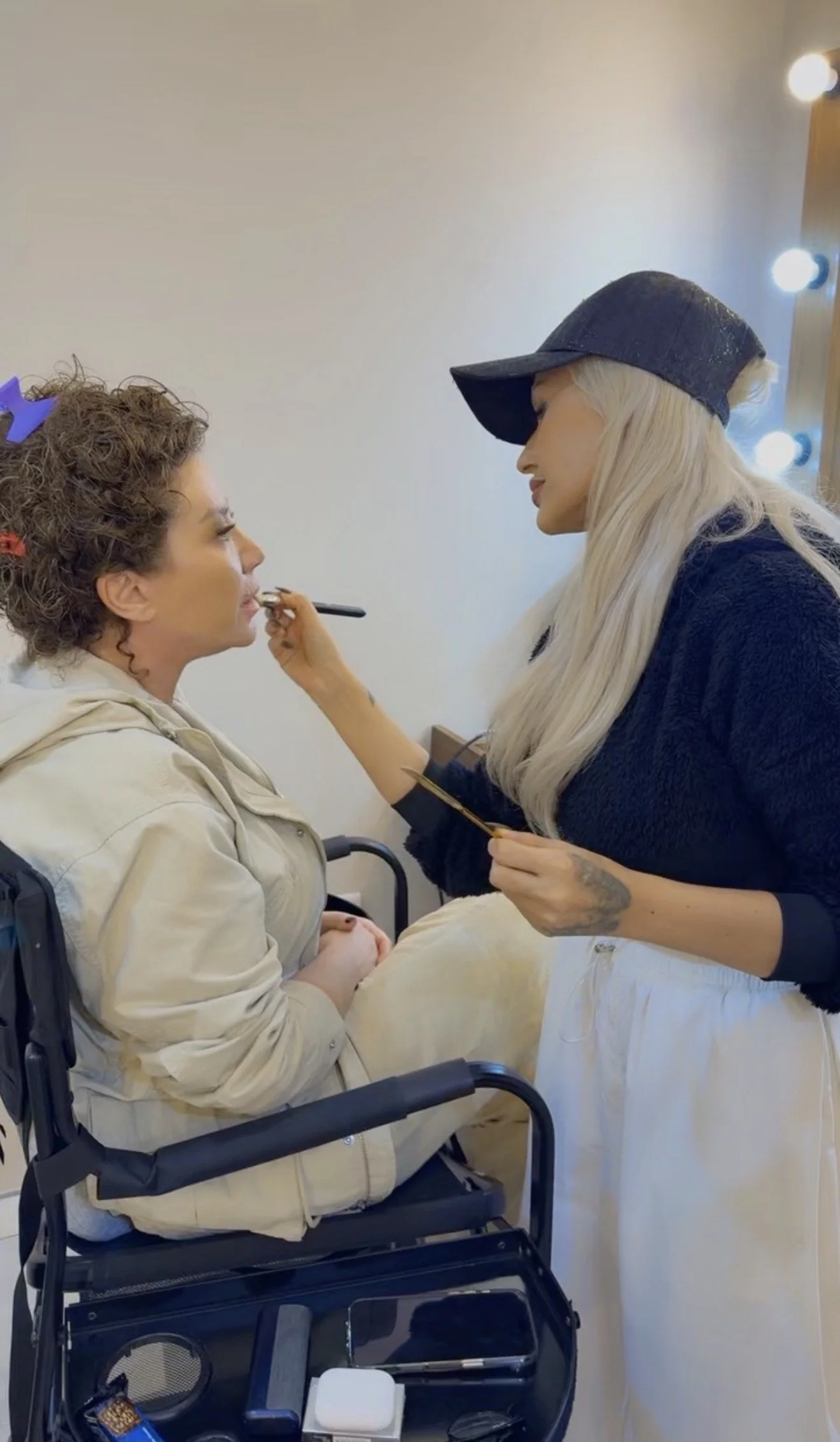 A makeup artist applying makeup to a woman in a wheelchair with curlers in her hair, in front of a mirror with lights.