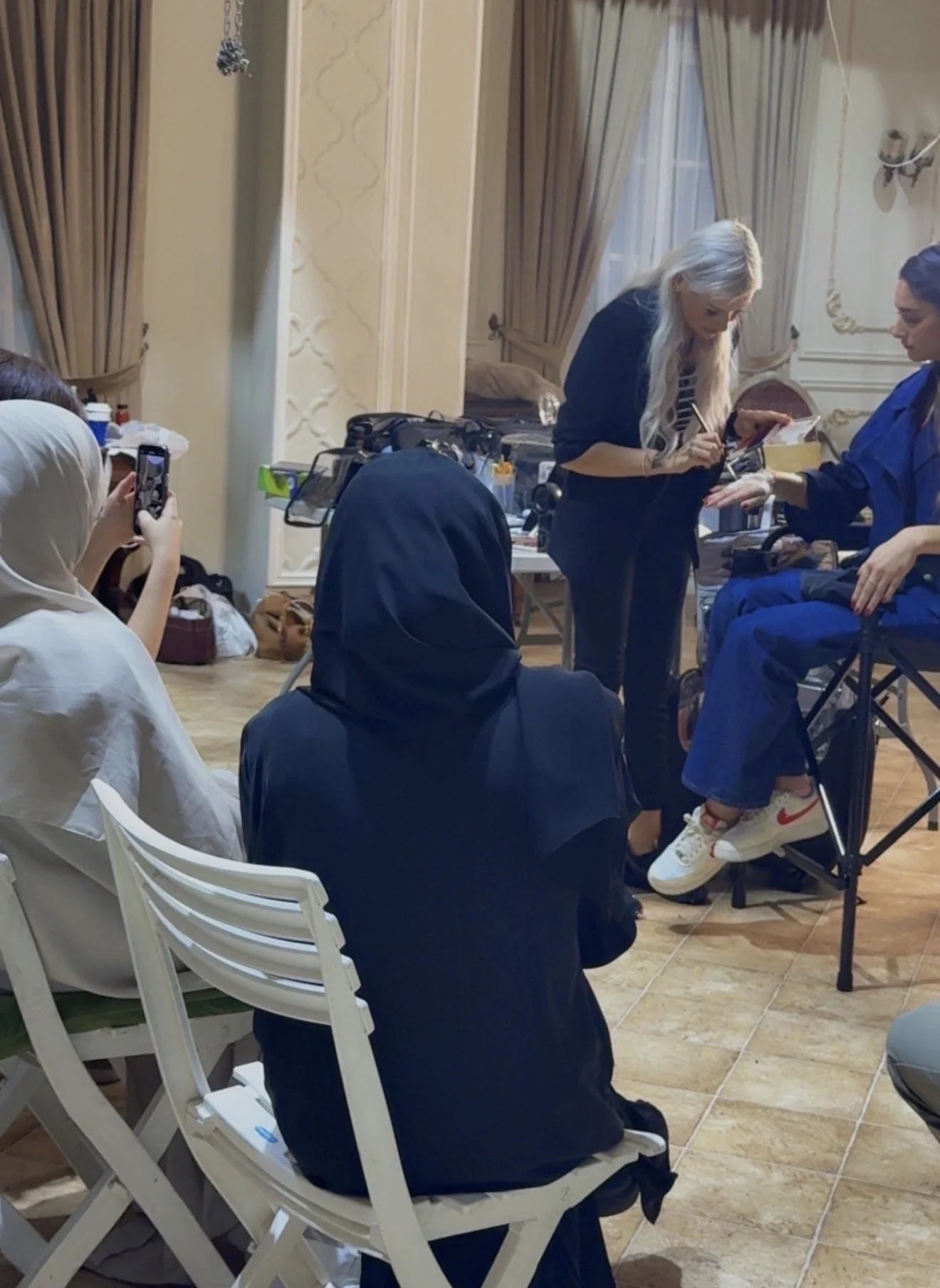 A woman with long blonde hair signing an autograph for a person seated in a wheelchair. Several people are watching, one person is taking a photo with a phone. The setting appears to be indoors with elegant curtains and decor.