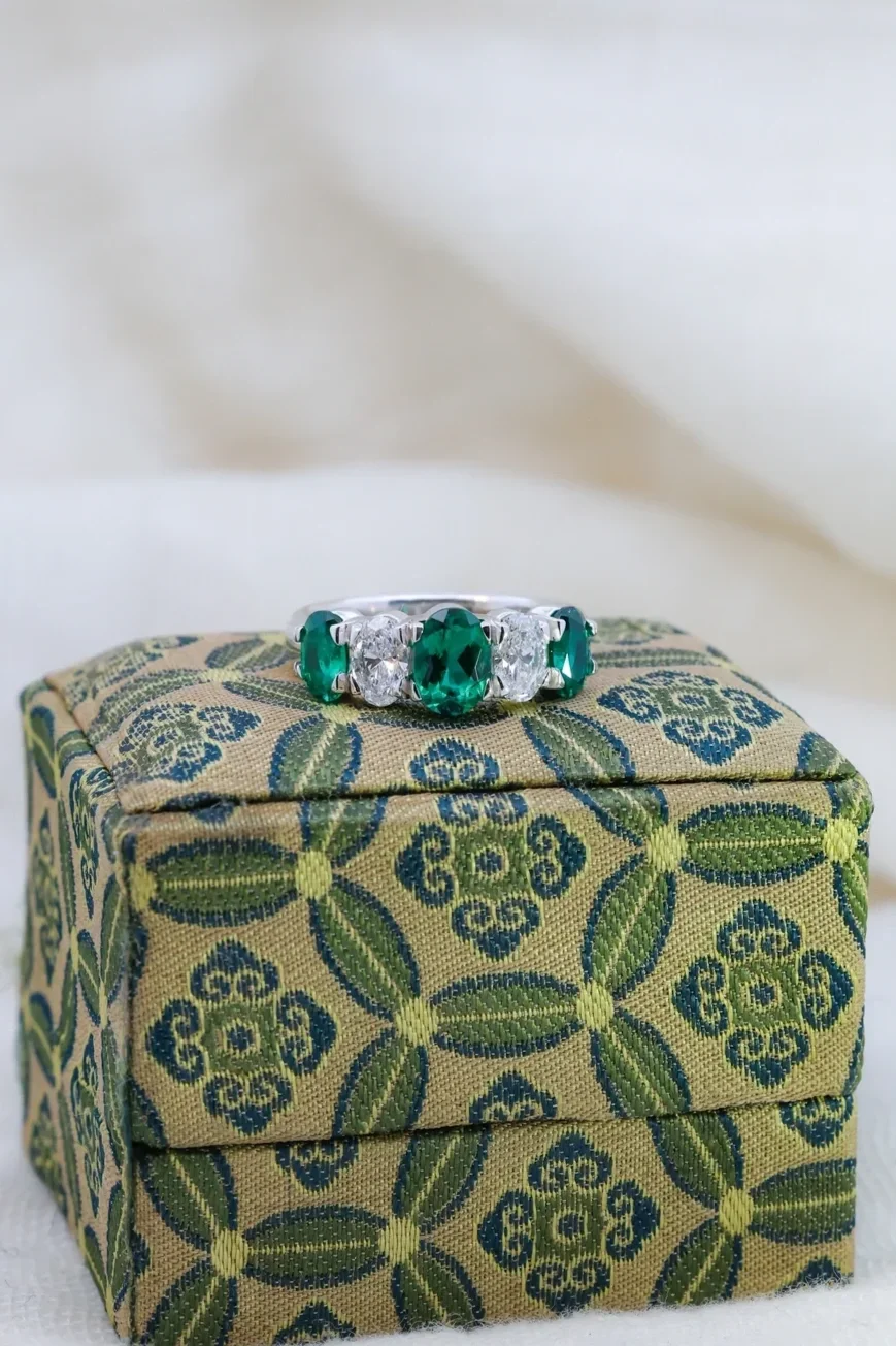 Custom white gold five stone emerald ring with alternating oval emeralds and diamonds placed on a green fabric ring box