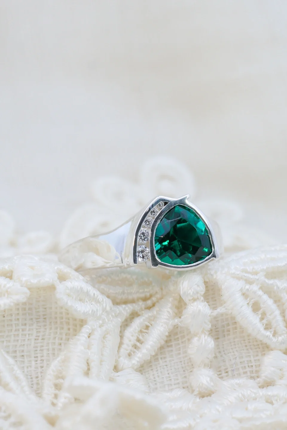 Custom white gold emerald trillion cut ring with offset diamond row placed on white lace