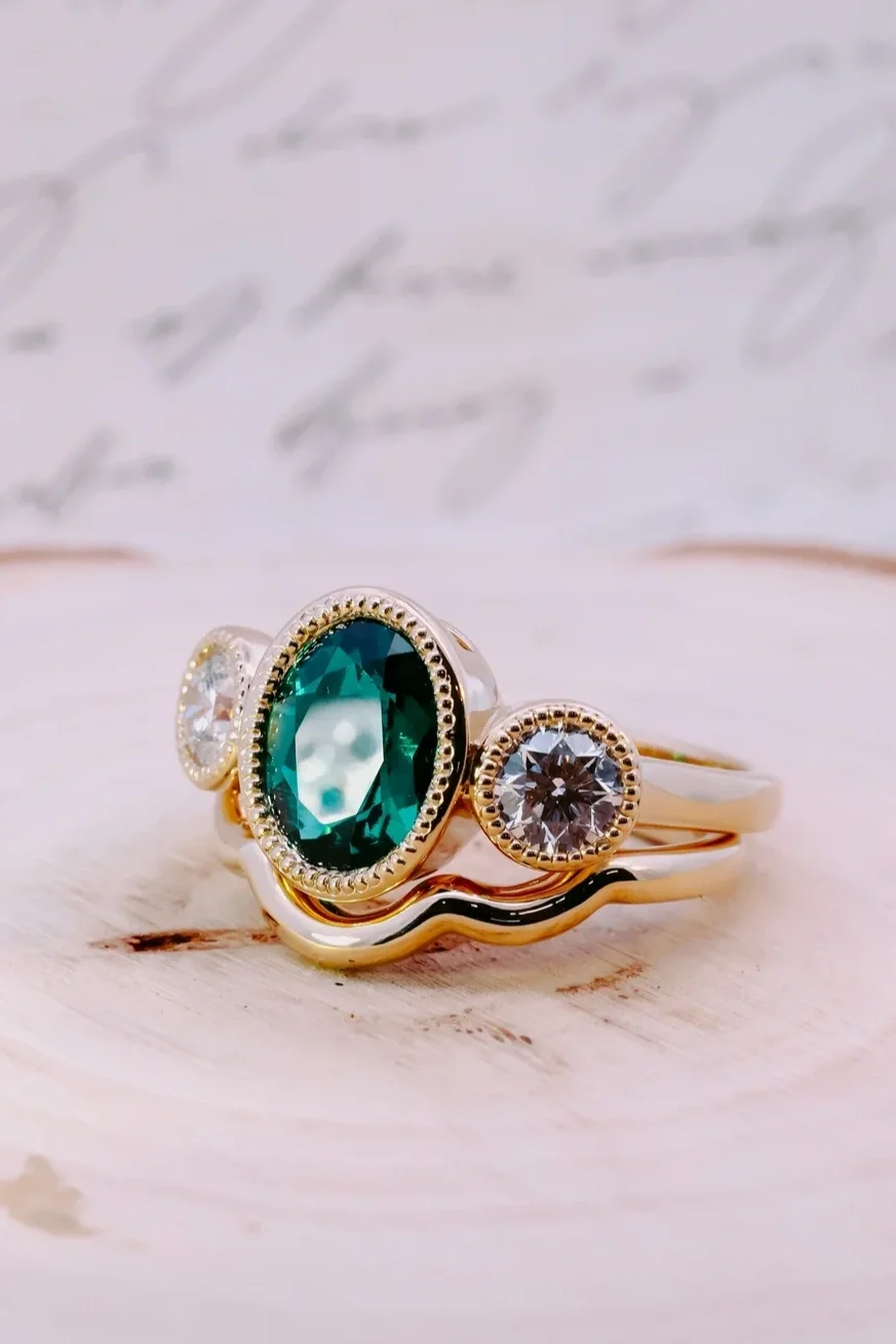 Custom vintage bezel set emerald three stone ring in yellow gold with oval emerald center stone and diamond side stones and matching yellow gold band